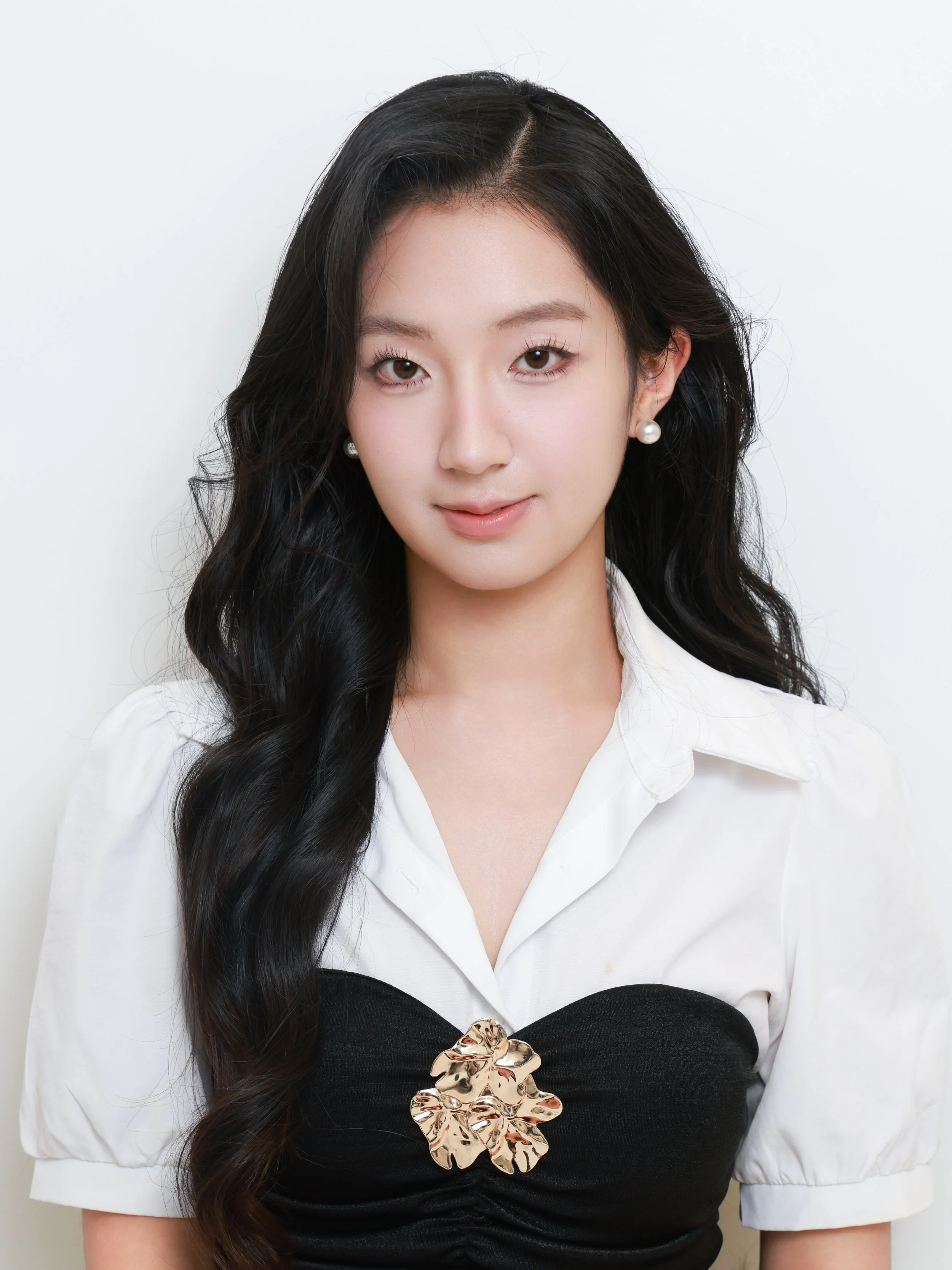A woman with long black wavy hair wearing a white blouse, black top, and pearl earrings, with a gold decorative brooch on her top, against a plain white background.