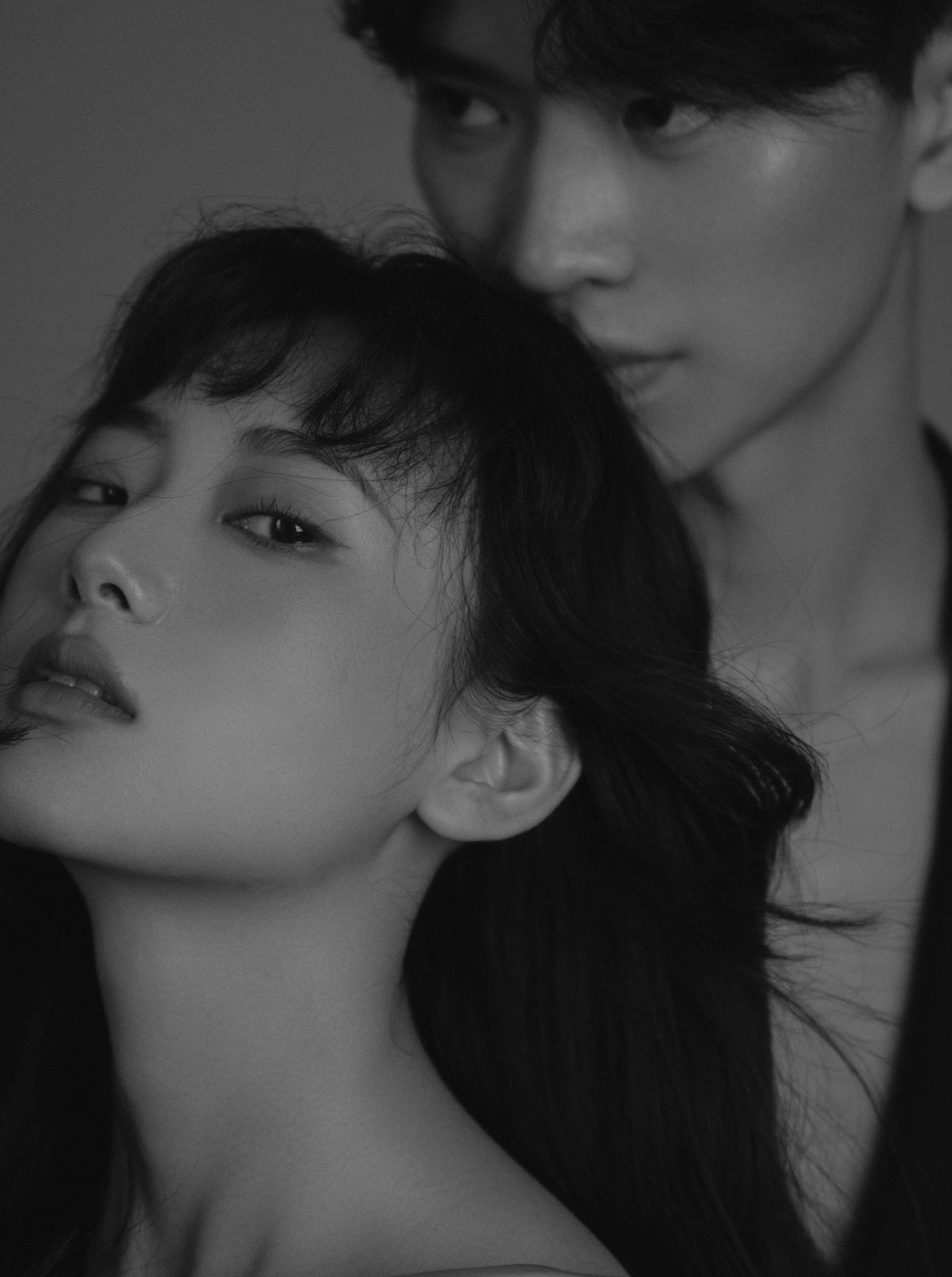 Black and white close-up photograph of a woman with dark hair and a man with dark hair in the background, both with intense expressions.