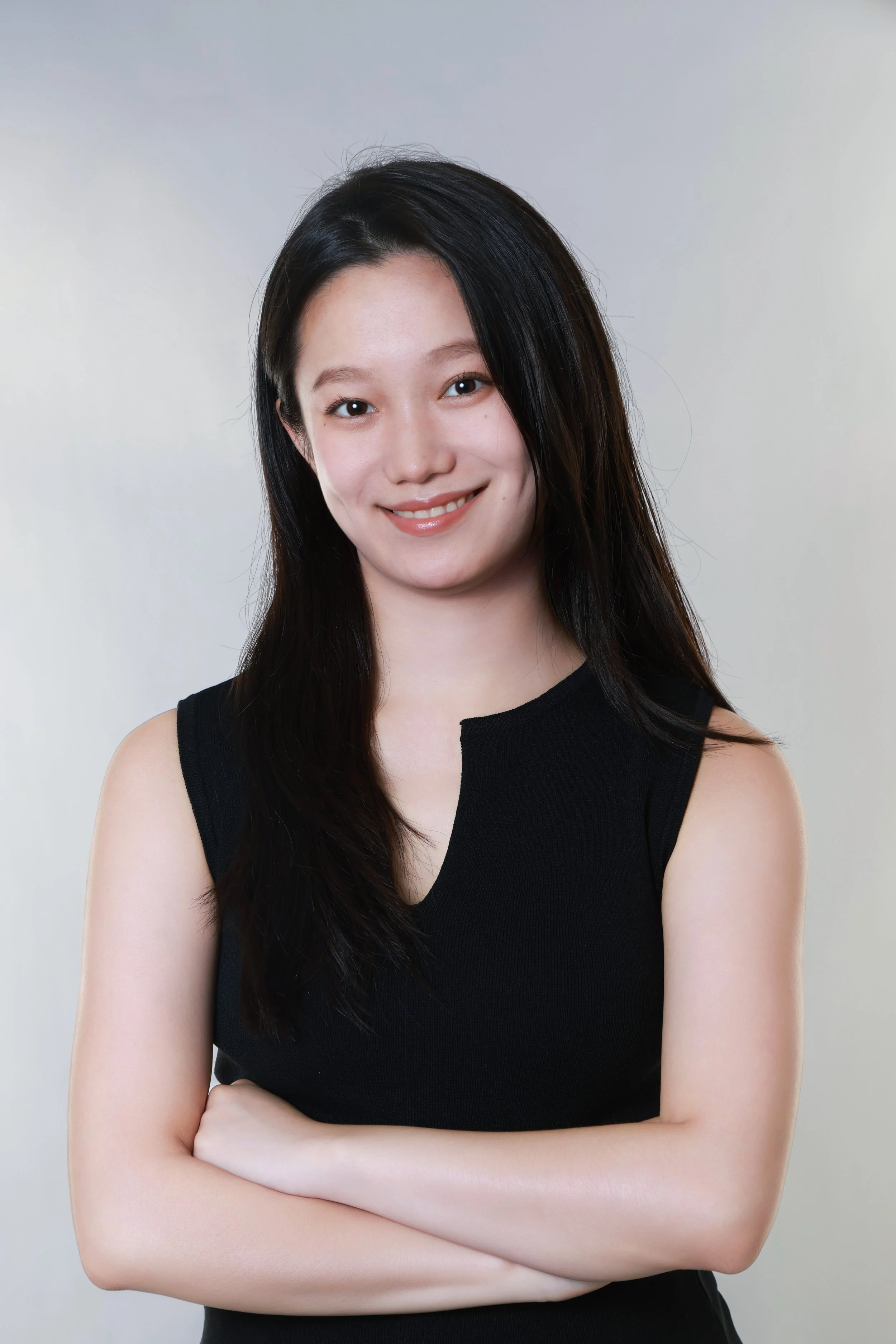 A young woman with long dark hair wearing a sleeveless black top, smiling with arms crossed, against a light gray background.