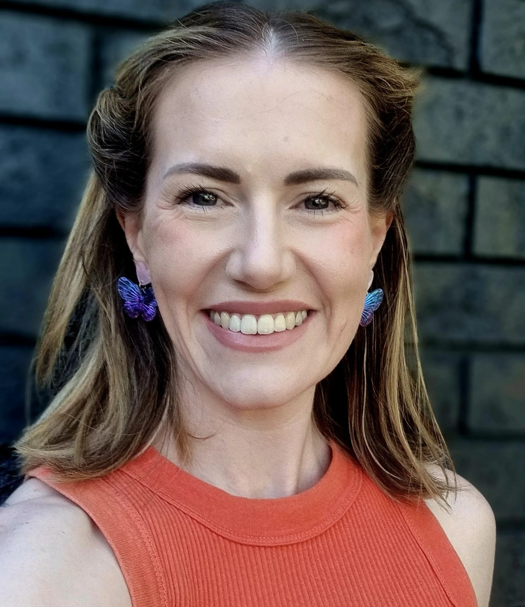 Close-up of a smiling woman with brown hair, wearing butterfly earrings and an orange sleeveless top, standing in front of a stone wall.