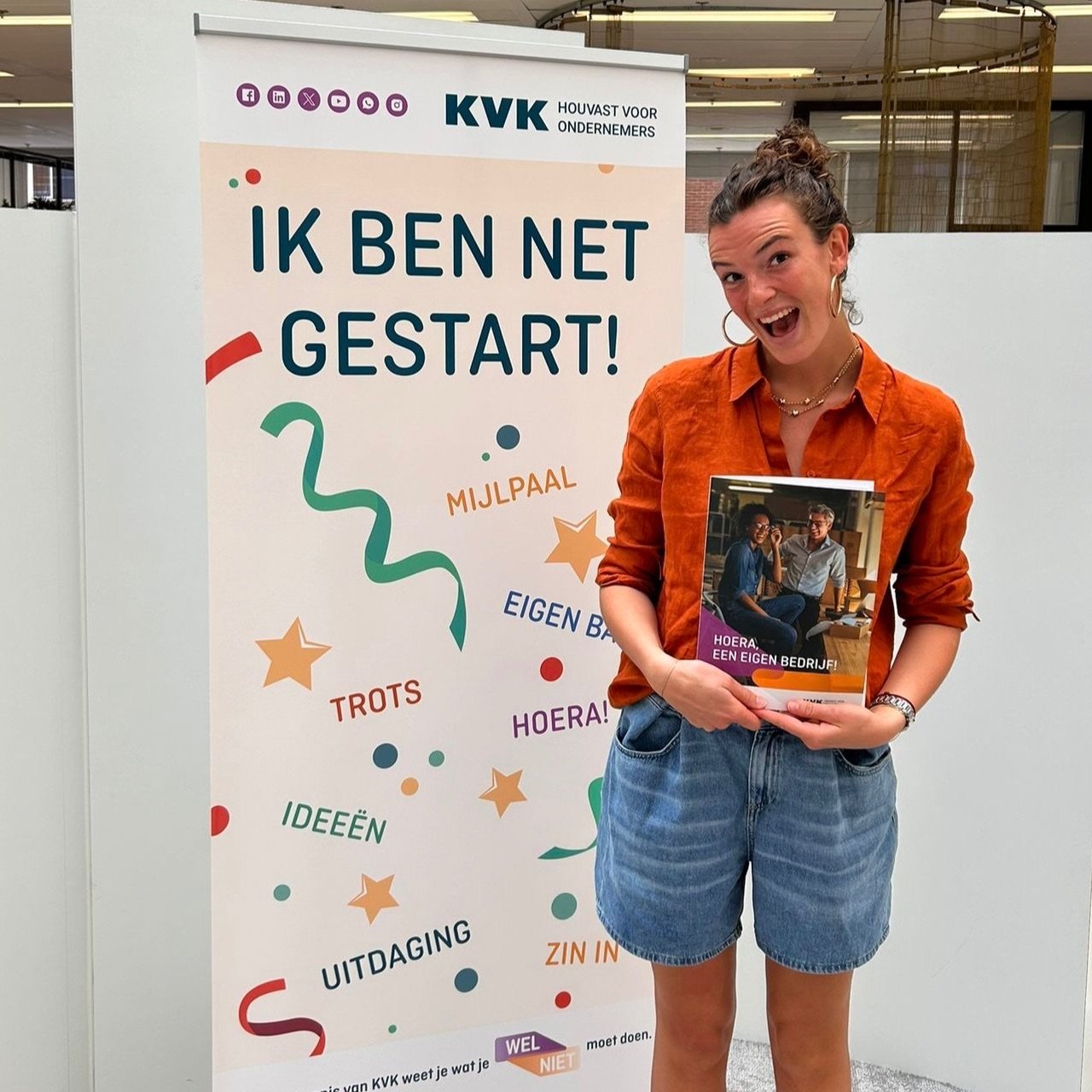 Vrouw staat naast een poster met de tekst 'IK BEN NET GESTART!' en andere woorden zoals 'MIJLPAA', 'EIGEN BED', 'TROTS', 'HOERA!', 'IDEEËN', 'UITDAGING', 'ZIN IN'. Ze houdt een brochure vast met een foto van twee gelukkige mensen en de tekst 'HOERA! EEN EIGEN BEDRIJF!'
