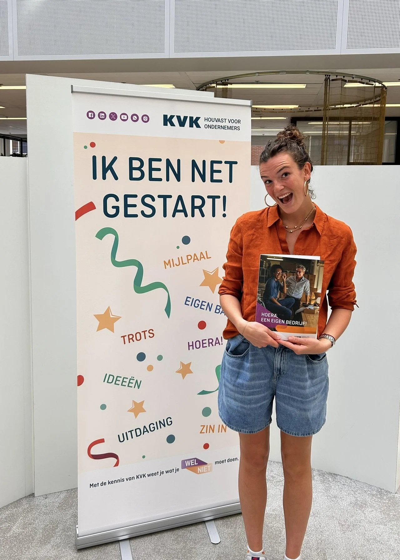 Een jonge vrouw staat naast een promotiebord van KVK en houdt een brochure vast. Ze draagt een oranje blouse en een denim rok, en lijkt enthousiast.