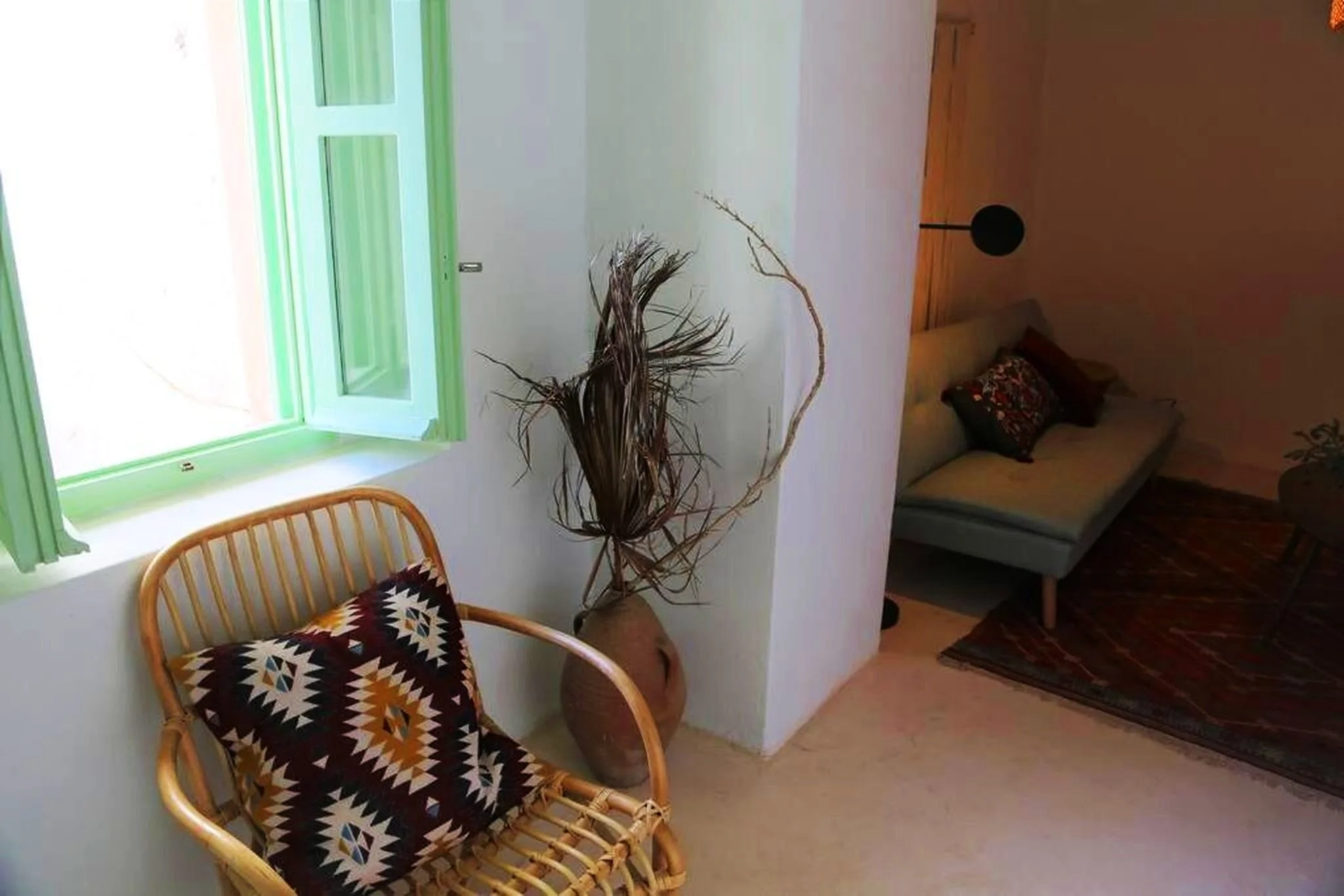 A cozy living room corner with a rattan chair and decorative pillow, a large ceramic vase with dried twigs, a green window with open shutters, a light-colored sofa with pillows, a hanging black lamp, a patterned rug, and potted plants.
