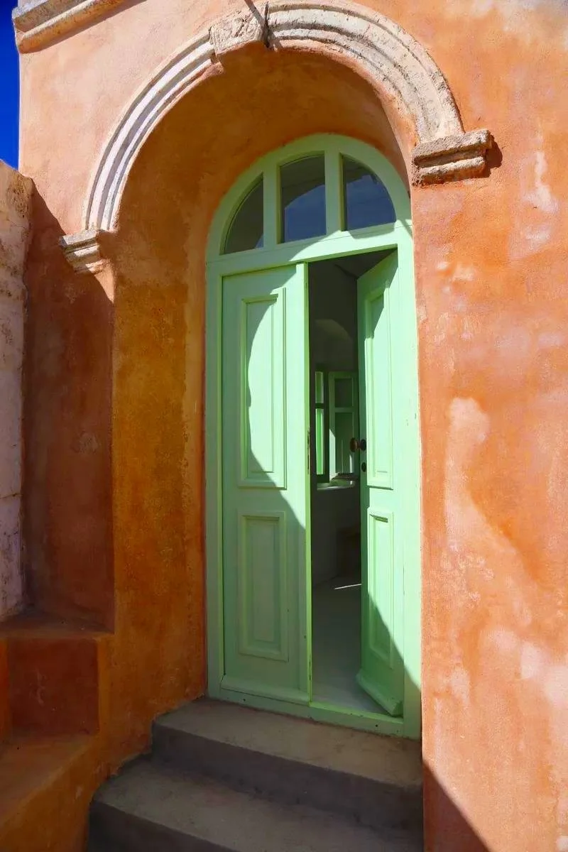 Open green door with an arched window above leading into a room with a mirror and sink, set in a peach-colored stucco wall with stairs to the left.