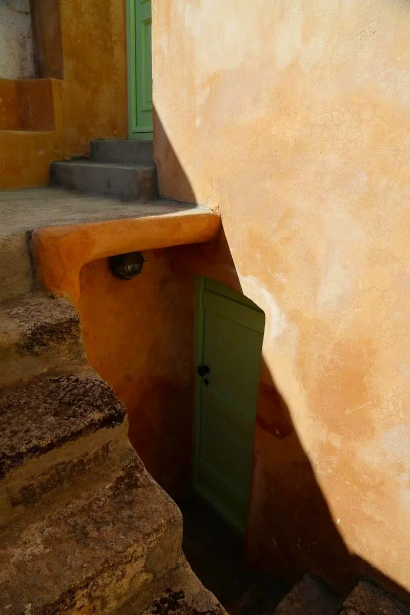 An outdoor staircase leading to a green door, with a small green door below, in a stucco wall with sunlight and shadow.