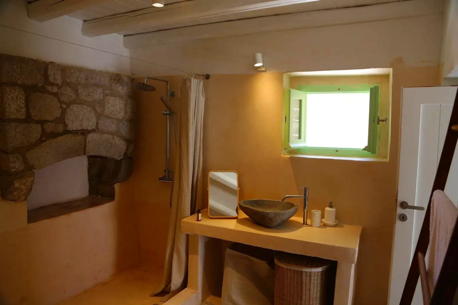 A rustic bathroom with a stone shower wall, a small window with green shutters, and a stone sink on a beige countertop.