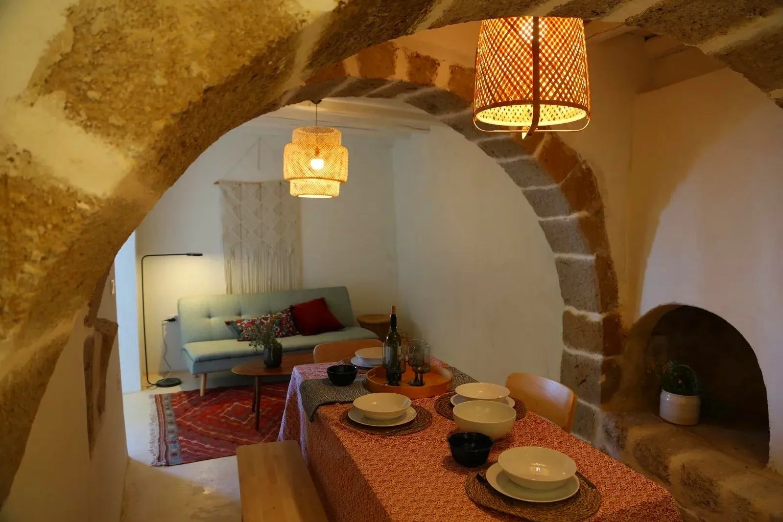 Dining area with a table set with bowls and glasses, overlooking a living room with a sofa, lamp, and decorative cushions, through stone archways.