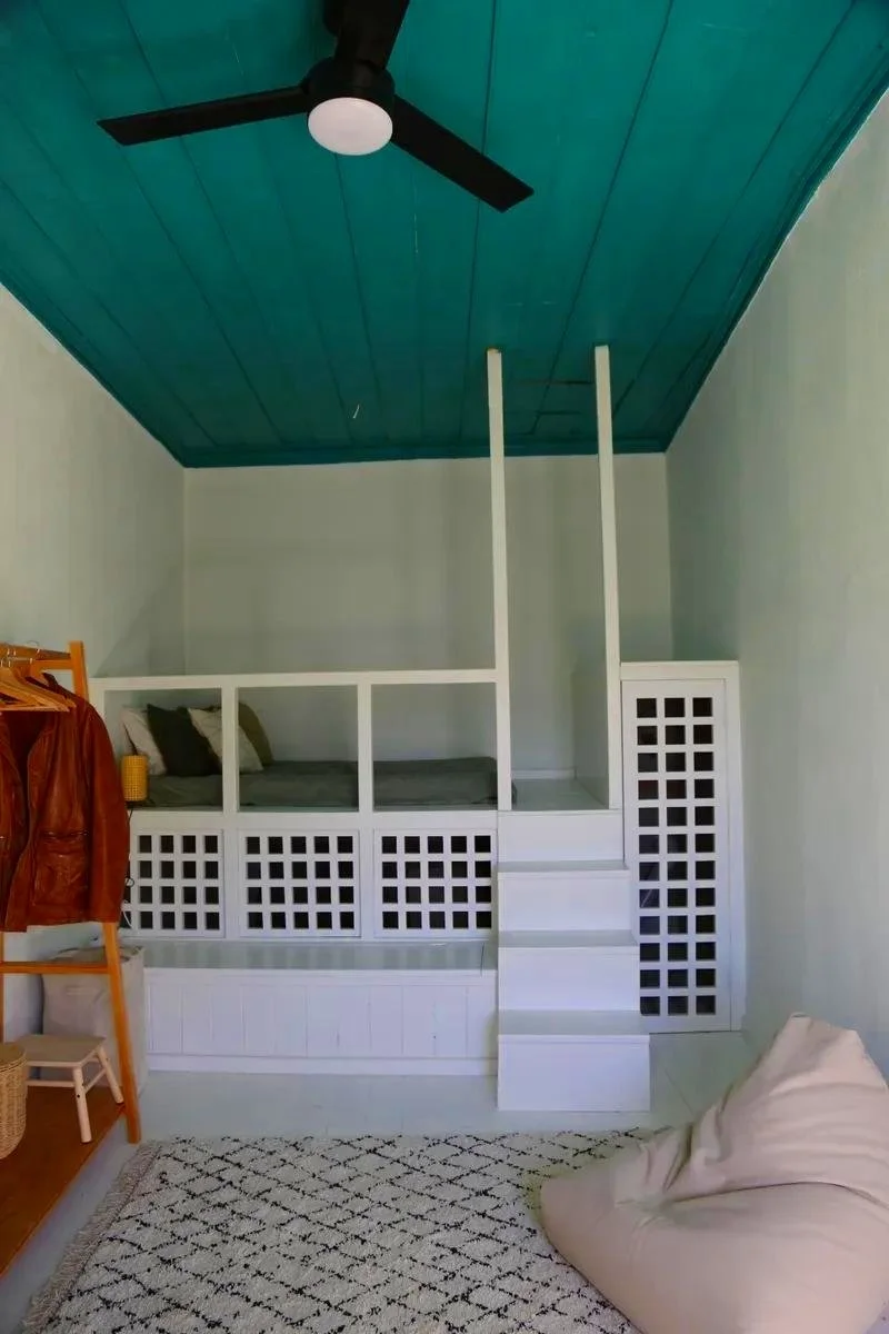 Small elevated bed with white lattice sides, green pillows, and a small set of stairs leading up to it, set against light-colored walls and a turquoise ceiling with a black ceiling fan.