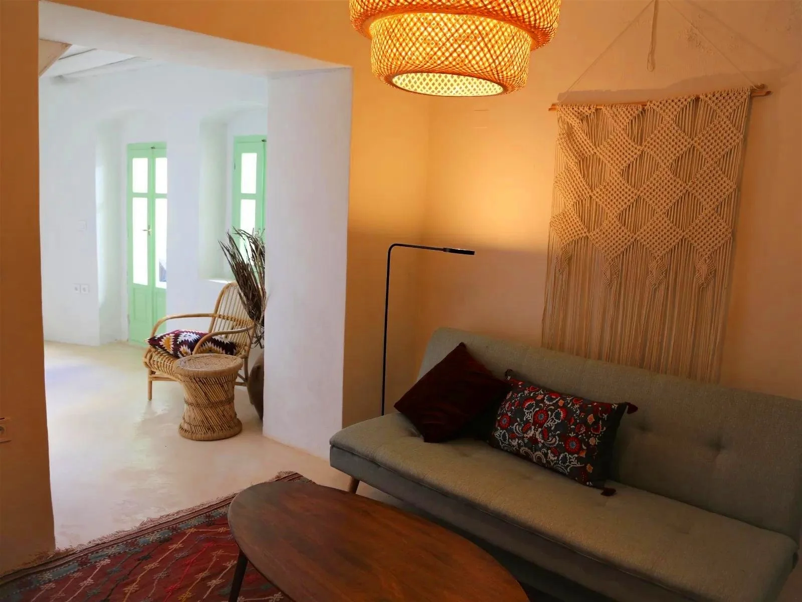 Cozy living room with a gray sofa, decorative pillows, warm lighting, woven wall hanging, and a wooden coffee table. Adjacent area features wicker chair with a pillow, dried plant decor, and green-framed windows and door.
