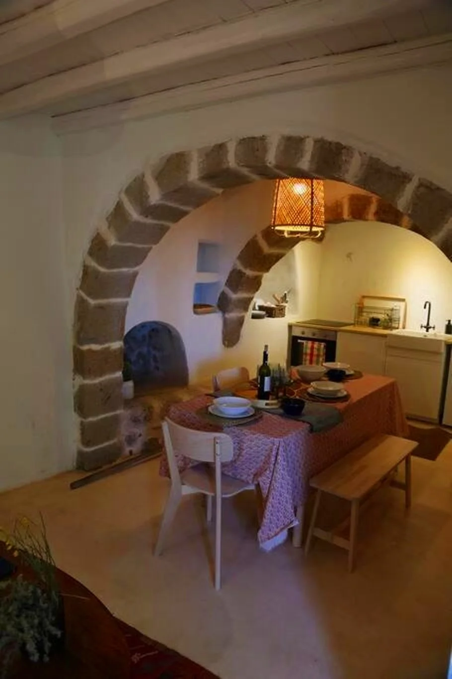 A cozy dining area with a wooden table covered with a red patterned tablecloth, set with white bowls, plates, wine bottle, and utensils. The room has rustic stone archways and warm ambient lighting, with a kitchen area in the background featuring a s