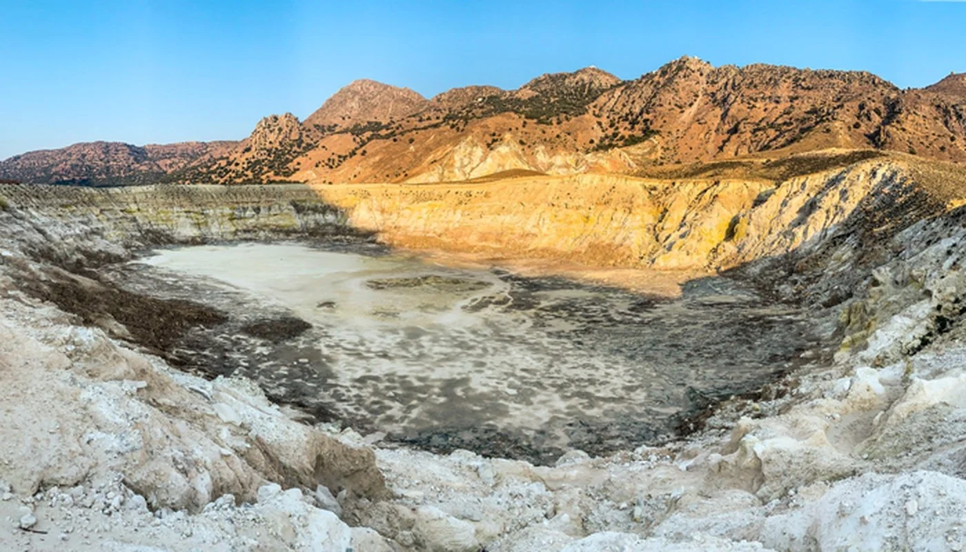A barren, crater-like depression in desert landscape with dry, cracked earth and colorful rolling hills in the background under a clear blue sky.
