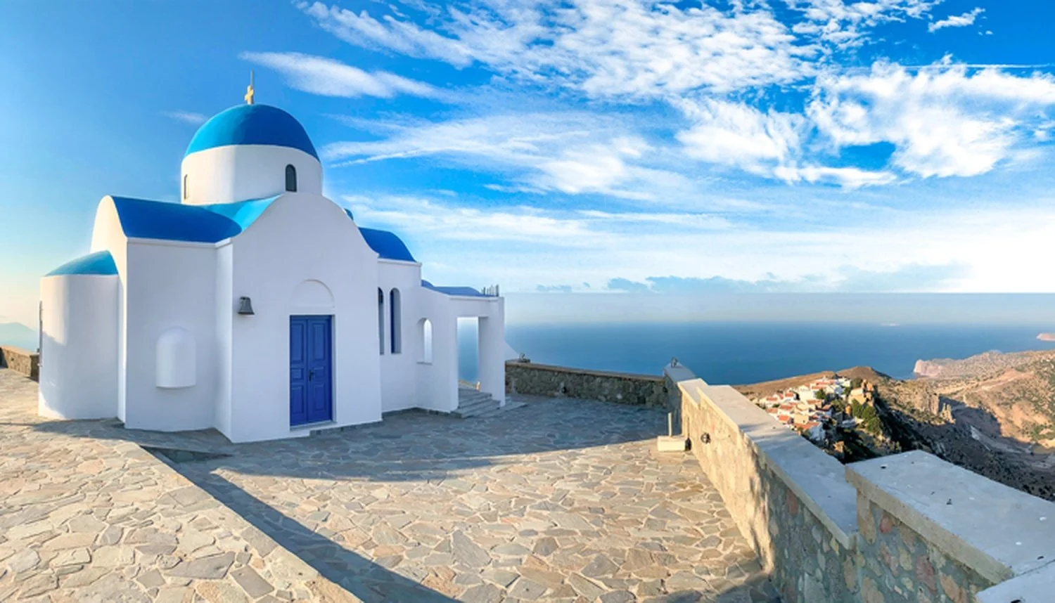 A white church with blue domes and door, overlooking an ocean and island landscape under a partly cloudy sky.