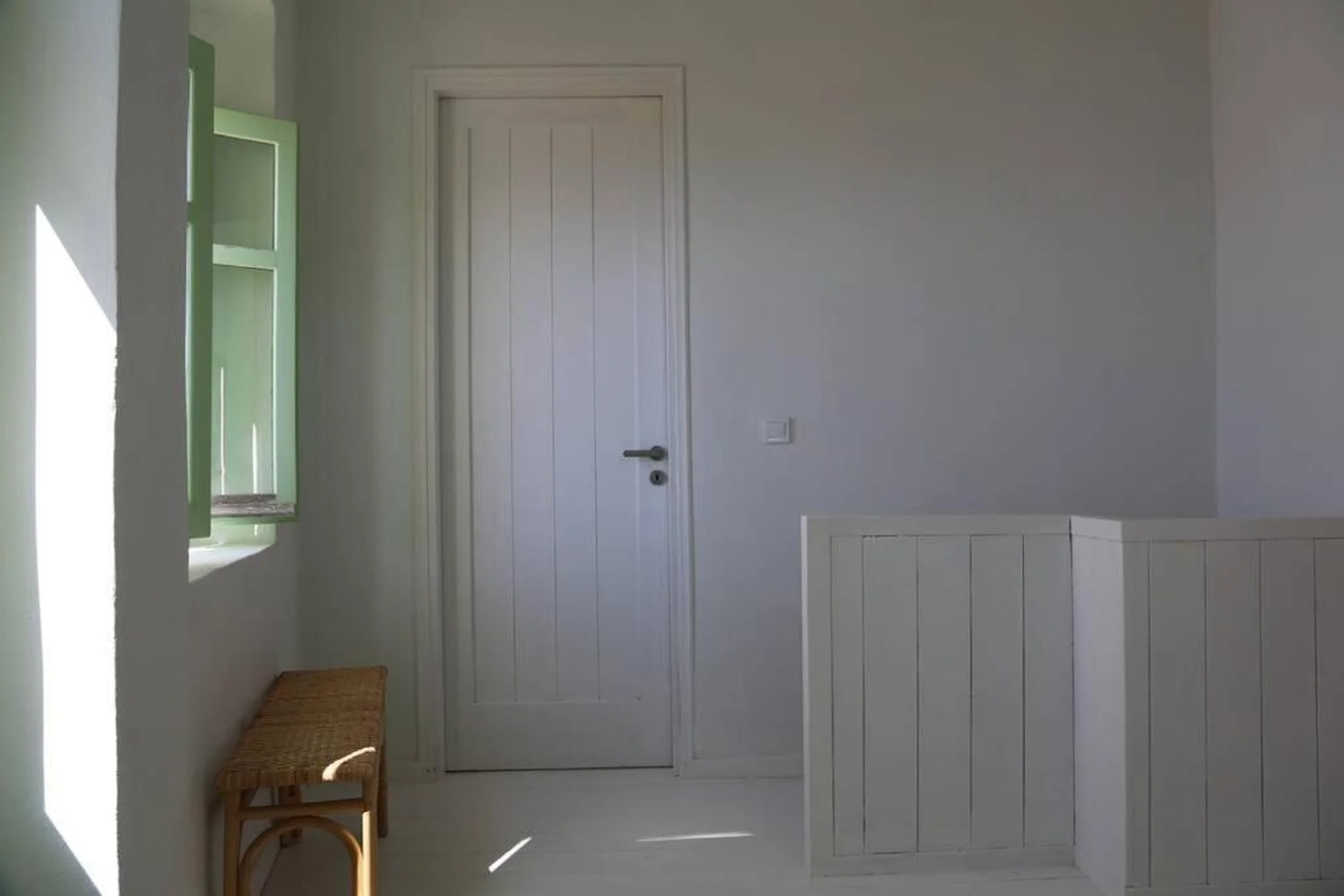 Interior of a small, minimalistic room with white walls, light green window shutters, a wooden bench, and a closed white door.