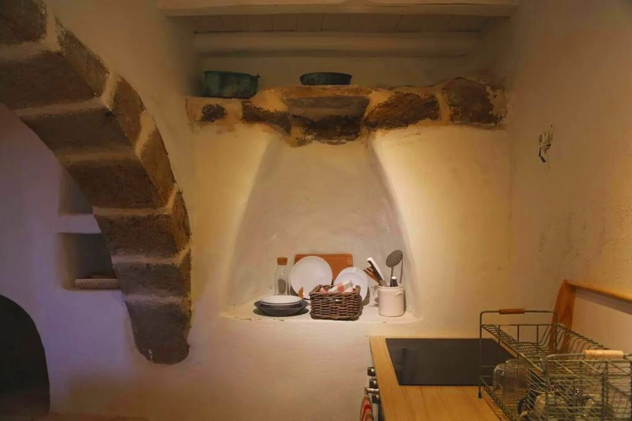 A small kitchen nook with a rustic stone arch and shelf, containing organized dishes and utensils, adjacent to a wooden countertop with a dish rack.