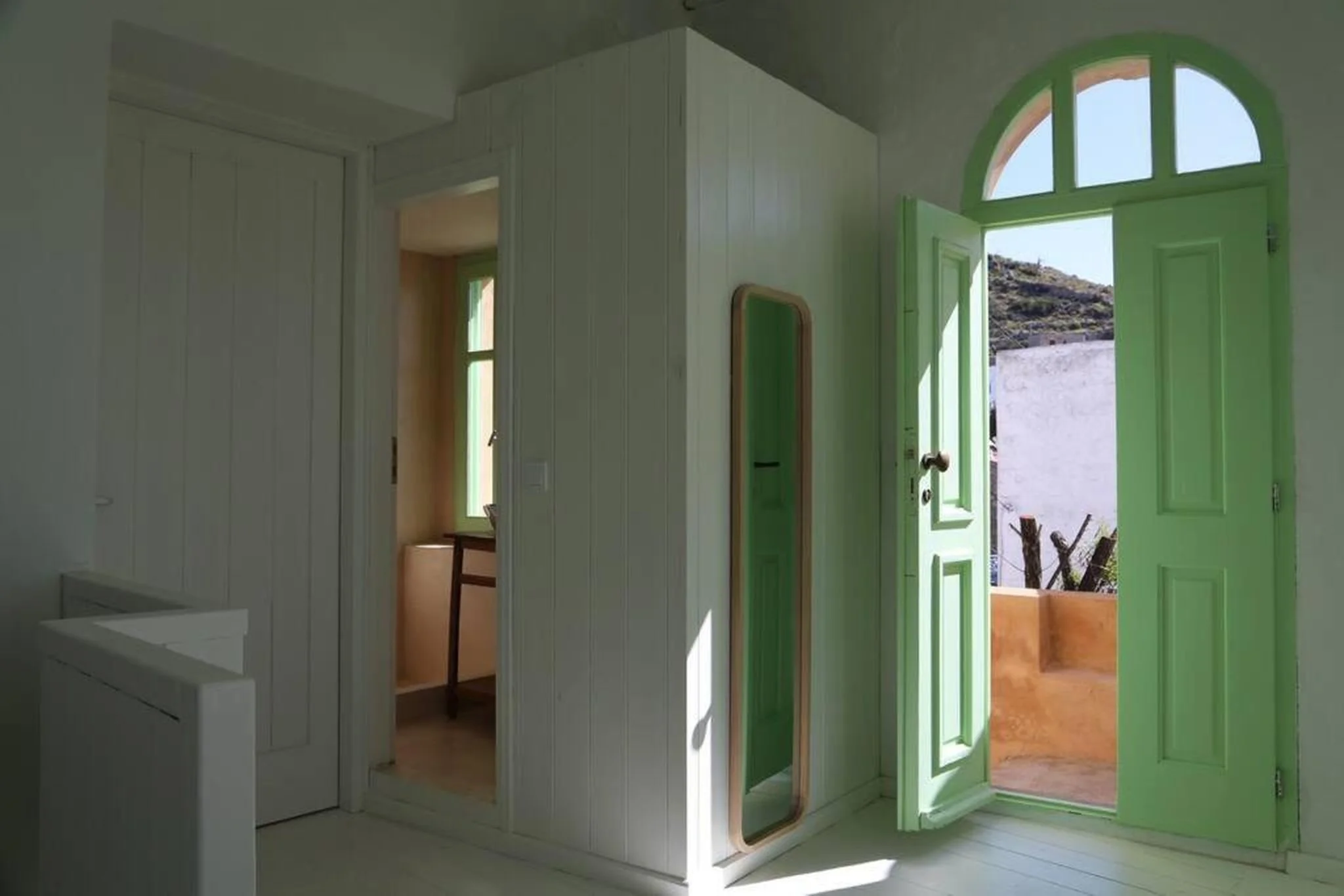 Interior of a house with a green front door open to an outdoor patio, and a mirror on the white wall.