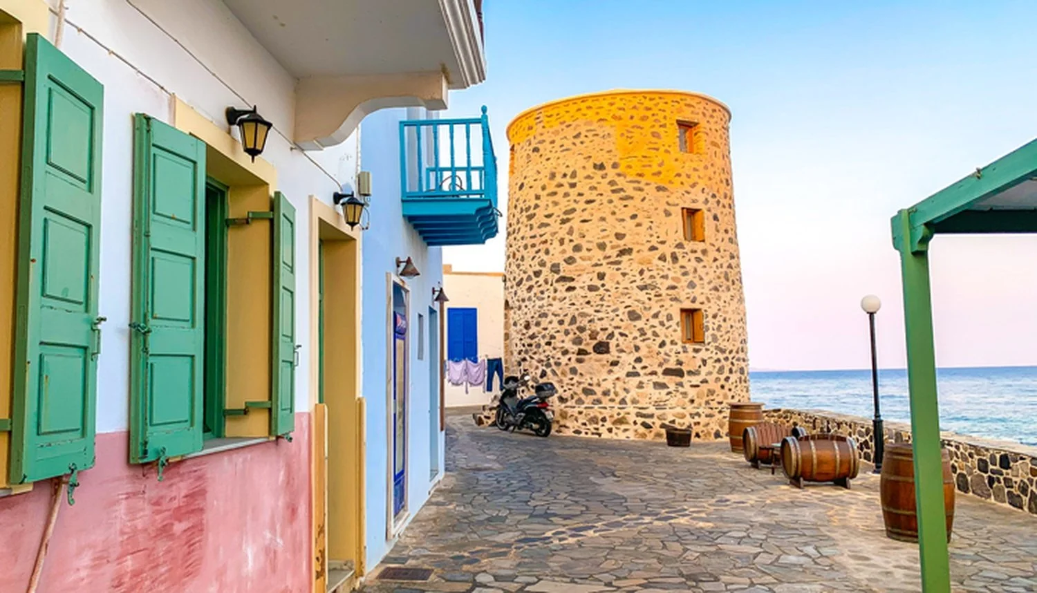 Colorful seaside scene with a stone tower, green window shutters, and a blue balcony under a clear sky.