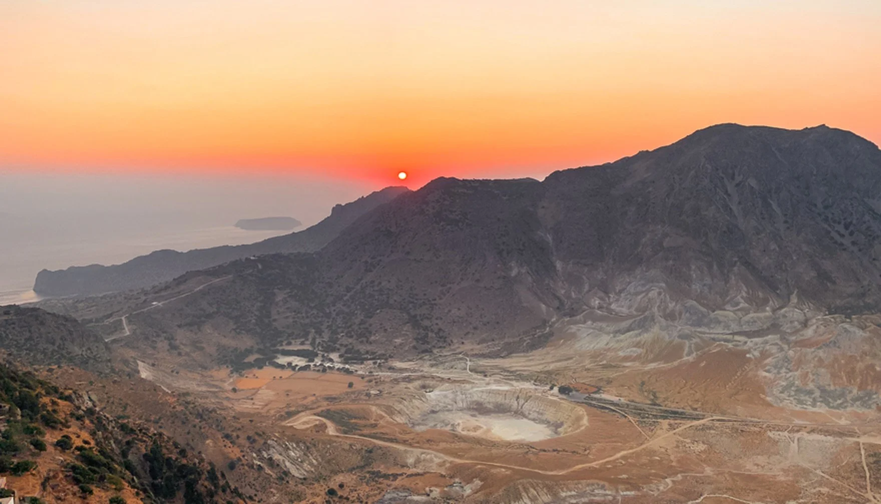 Sunset over rugged mountains near the coast with a crater in the foreground.