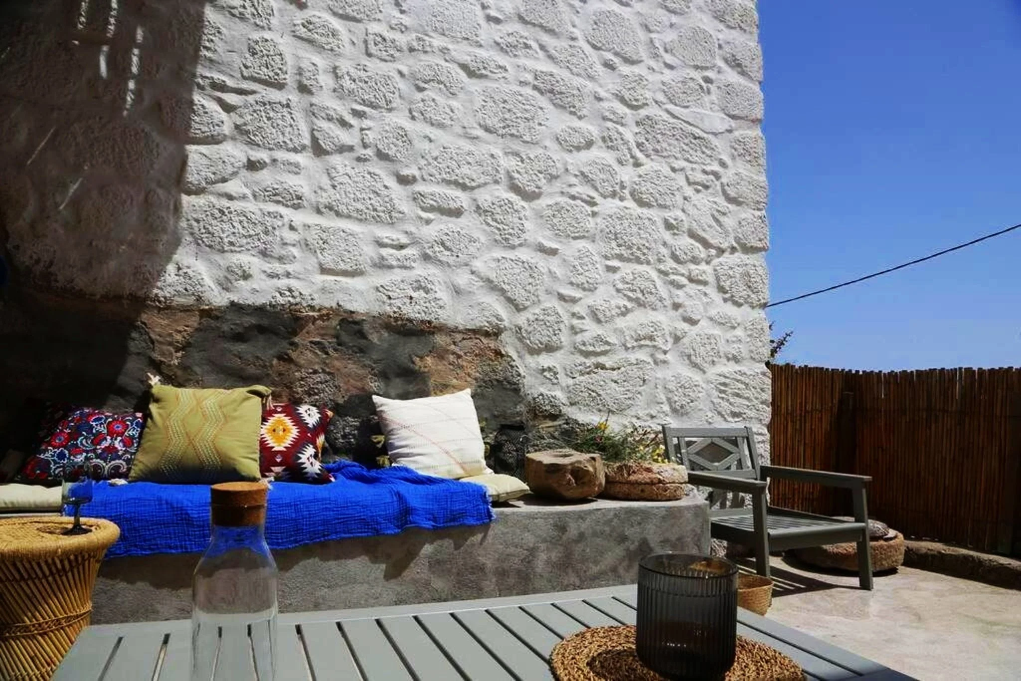 Outdoor patio with a stone wall, colorful cushions on a bench, a wooden chair, and a table with decorative items under a clear blue sky.