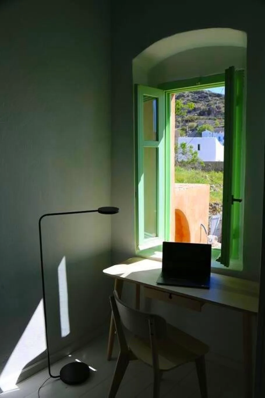 A simple interior room with a small desk near an open window with green shutters, a laptop on the desk, a modern black floor lamp, and sunlight casting shadows on the white walls and floor.