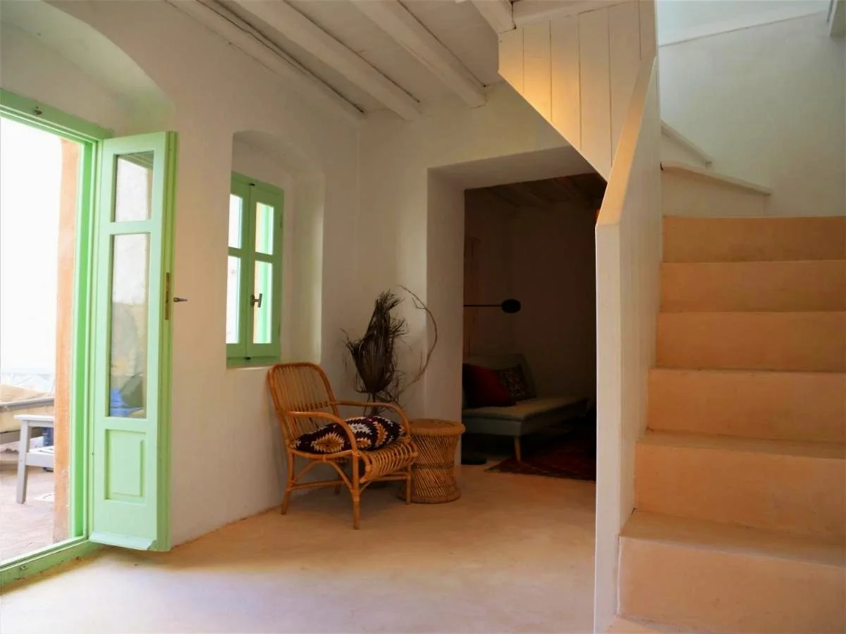 Interior of a house with white walls, light wooden floors, a green painted door and windows, a wicker chair with a patterned cushion, a small round wicker table, a built-in seating area with cushions under a wooden wall, and a staircase with light wo