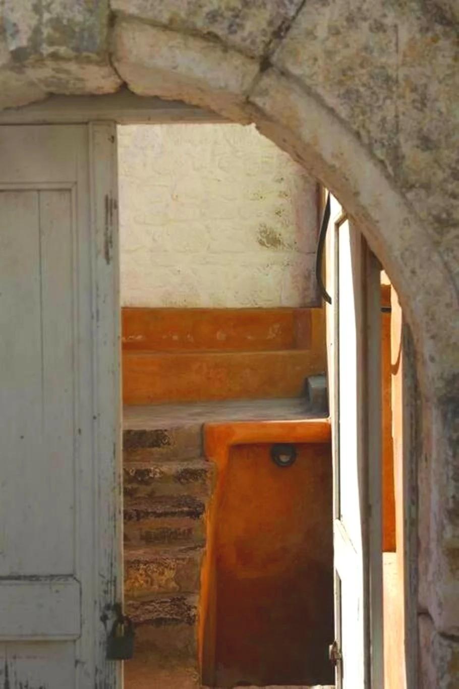 A stone archway leading to a small room with wooden stairs and a door, with stone walls and a wooden door frame.
