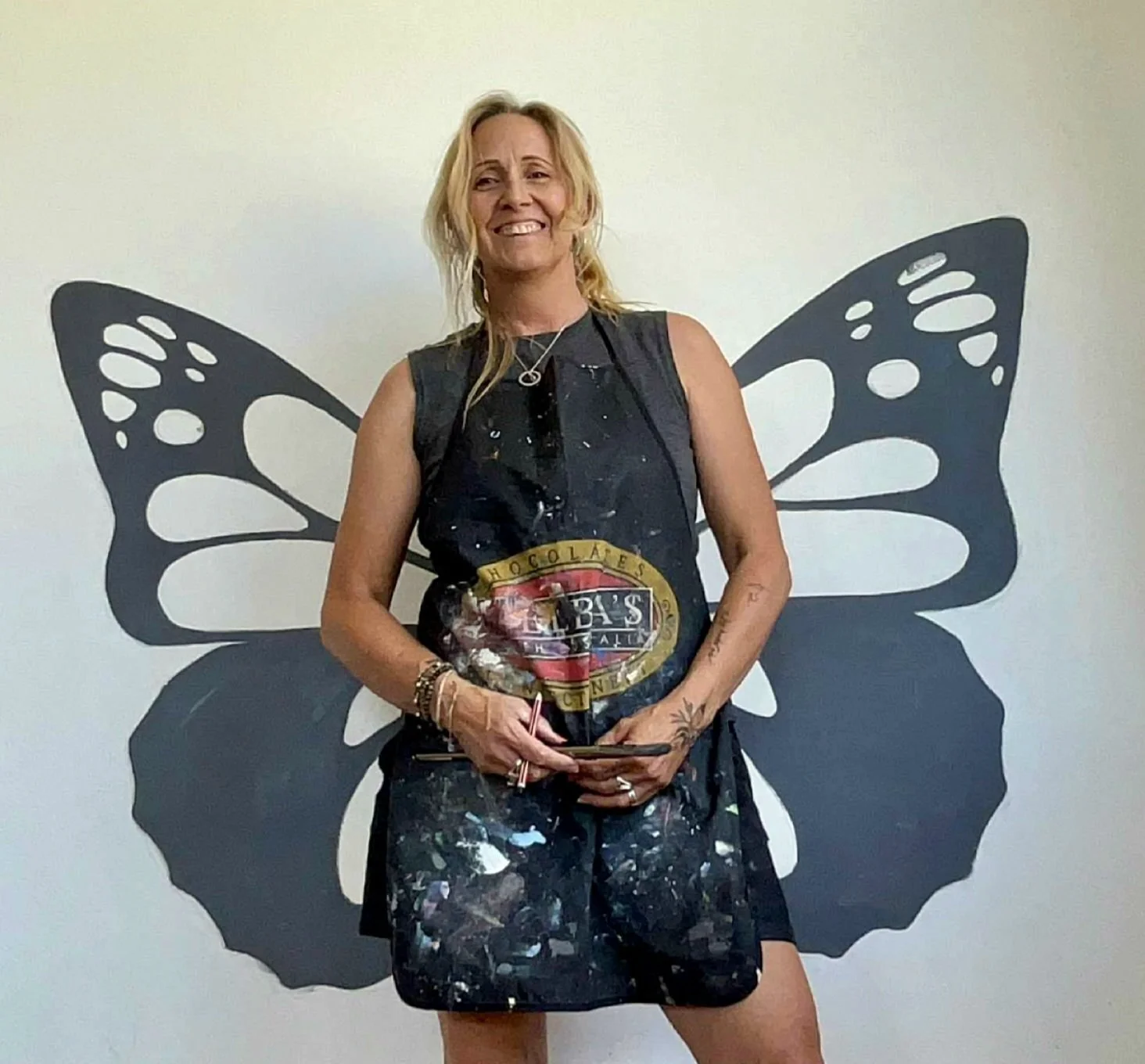 Woman with blonde hair smiling, posing in front of a butterfly-shaped wall art. She is wearing a black paint-splattered apron and holding a partially eaten chocolate bar and a smartphone.