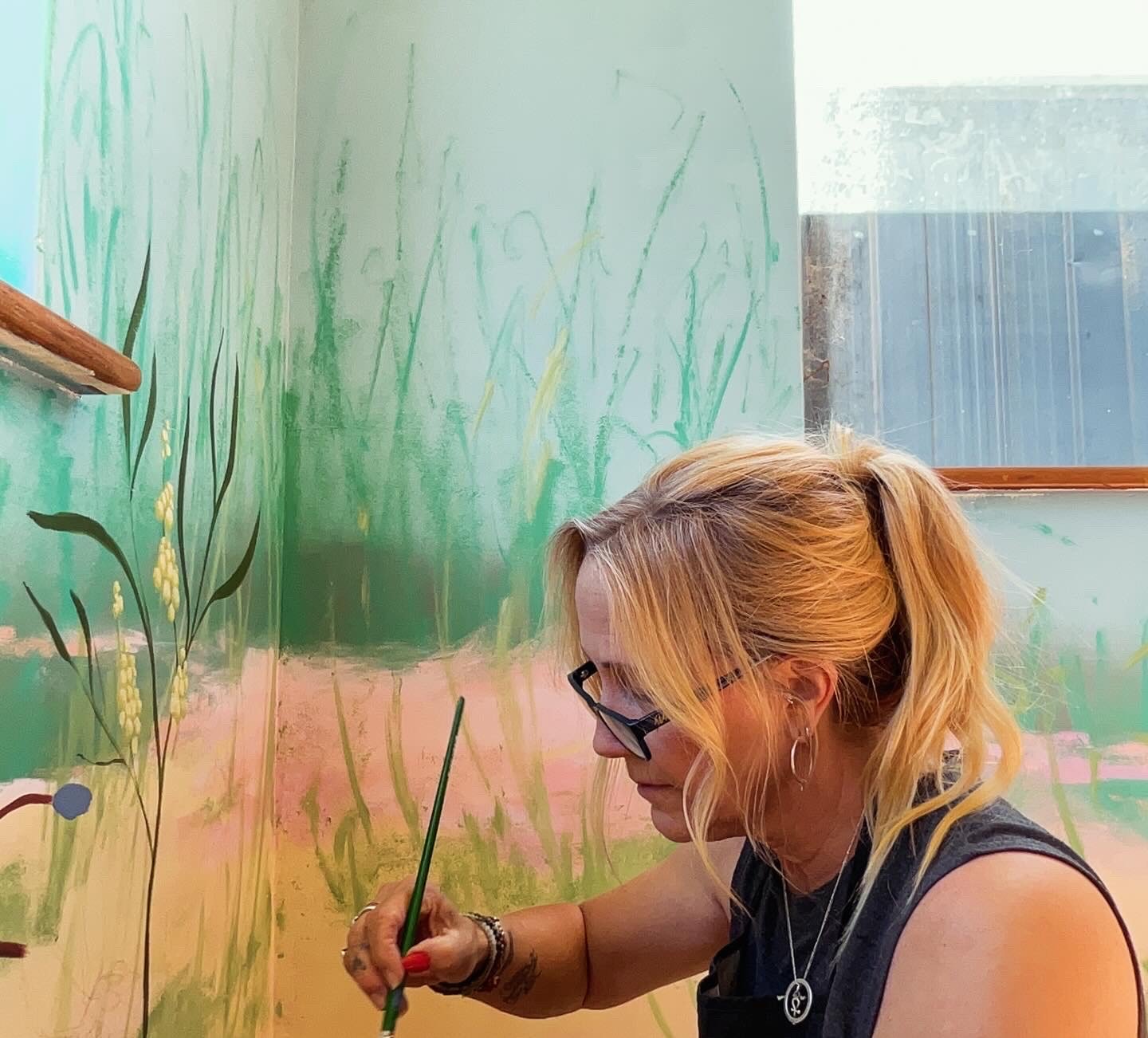 Woman with blonde hair, glasses, and earrings painting a mural of grass, flowers, and sky on a wall.