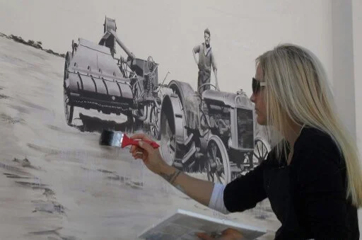 A woman with blonde hair and sunglasses painting a black and white mural of a vintage tractor on a wall.