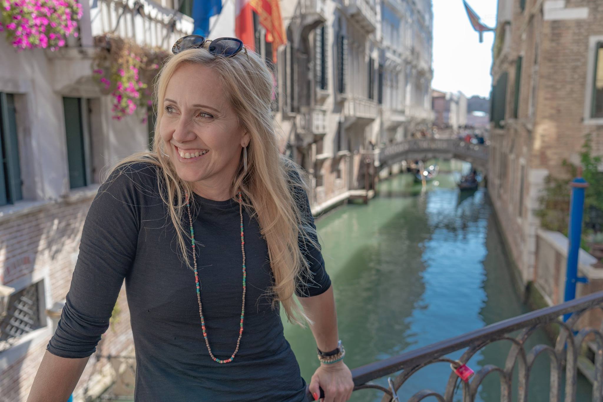 A smiling woman with blonde hair, wearing sunglasses on her head, a dark long-sleeve shirt, a colorful beaded necklace, and bracelets, stands on a bridge over a canal in Venice, Italy, with historic buildings and a small stone bridge in the background.