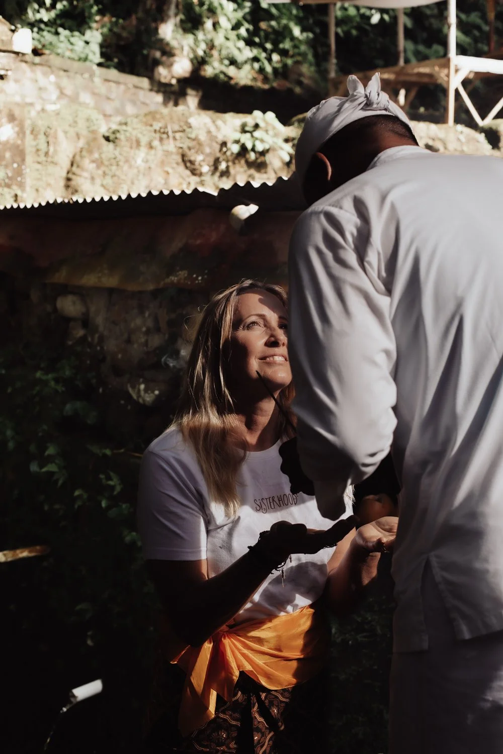 A woman with light brown hair smiling and looking up at a man in a white chef's coat and white headwrap, who is leaning over her. The woman is wearing a white t-shirt with the word "Sisterhood" on it, black gloves, and an orange sash. They are outdoors with trees, rocks, and some structures in the background.