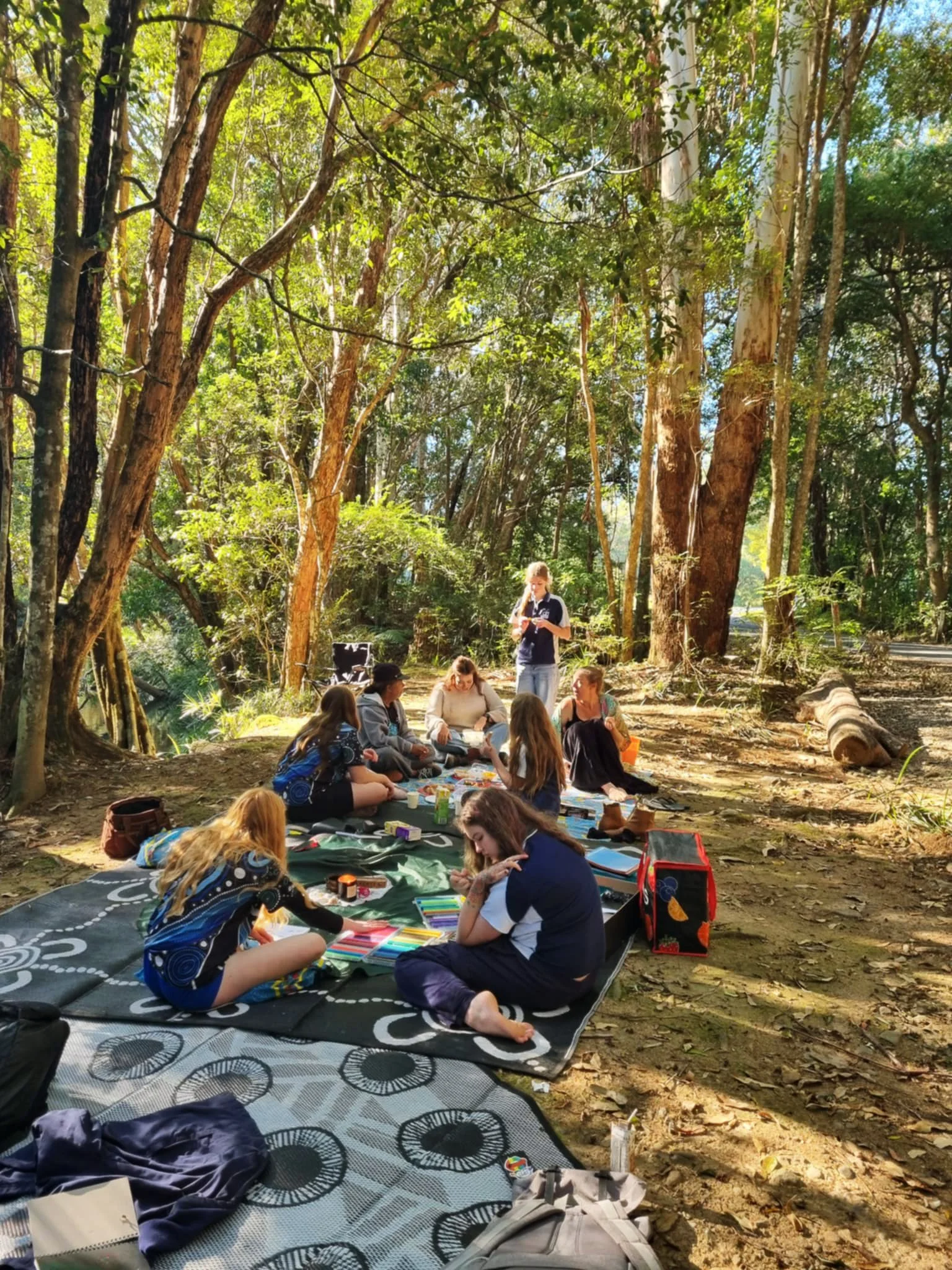 Group of children and adults sitting on mats in a forest, engaging in arts and crafts activities, with trees and sunlight overhead.