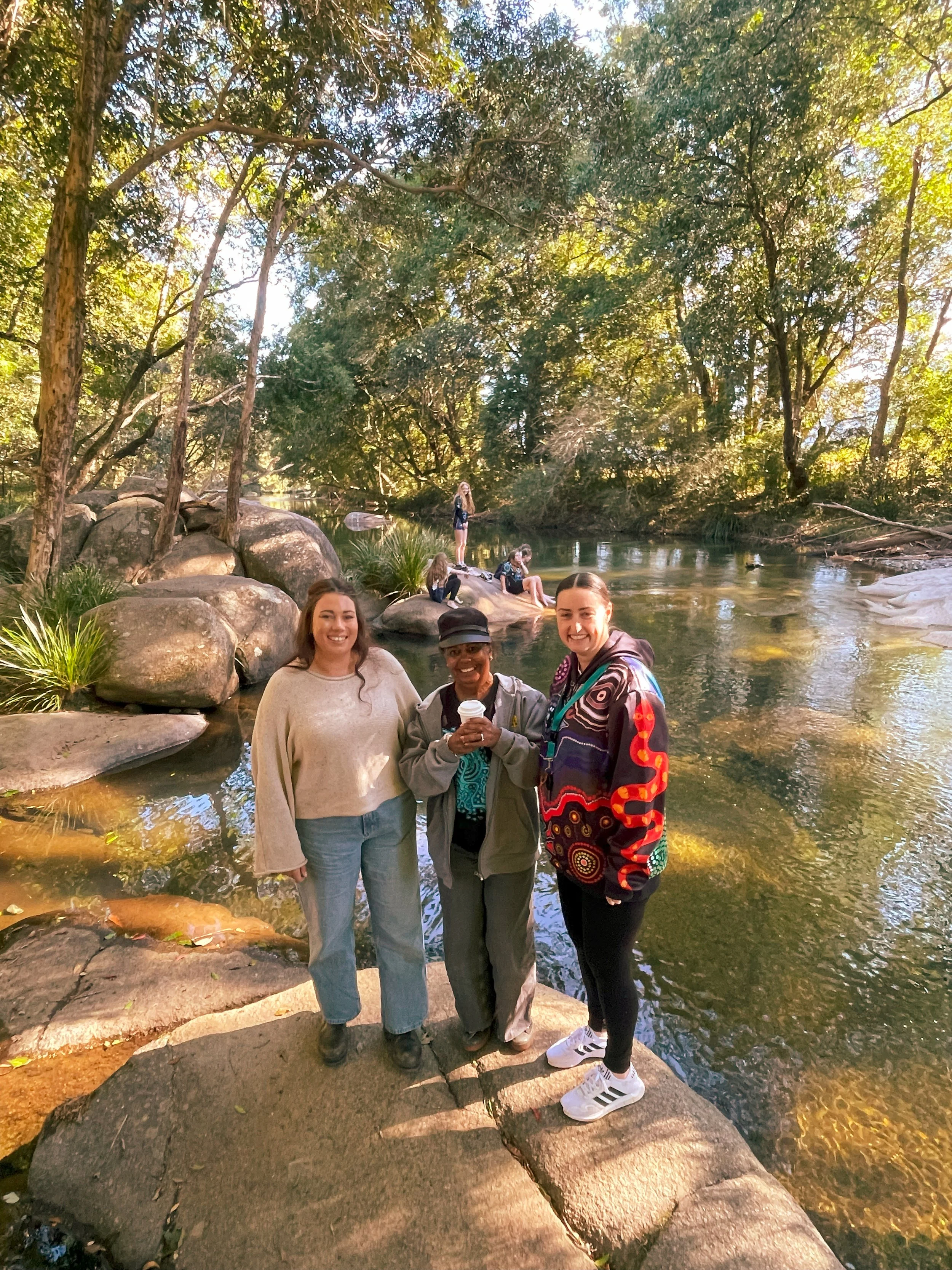 Three women standing on a rock by a river, surrounded by trees, with several people in the background enjoying the outdoor setting.