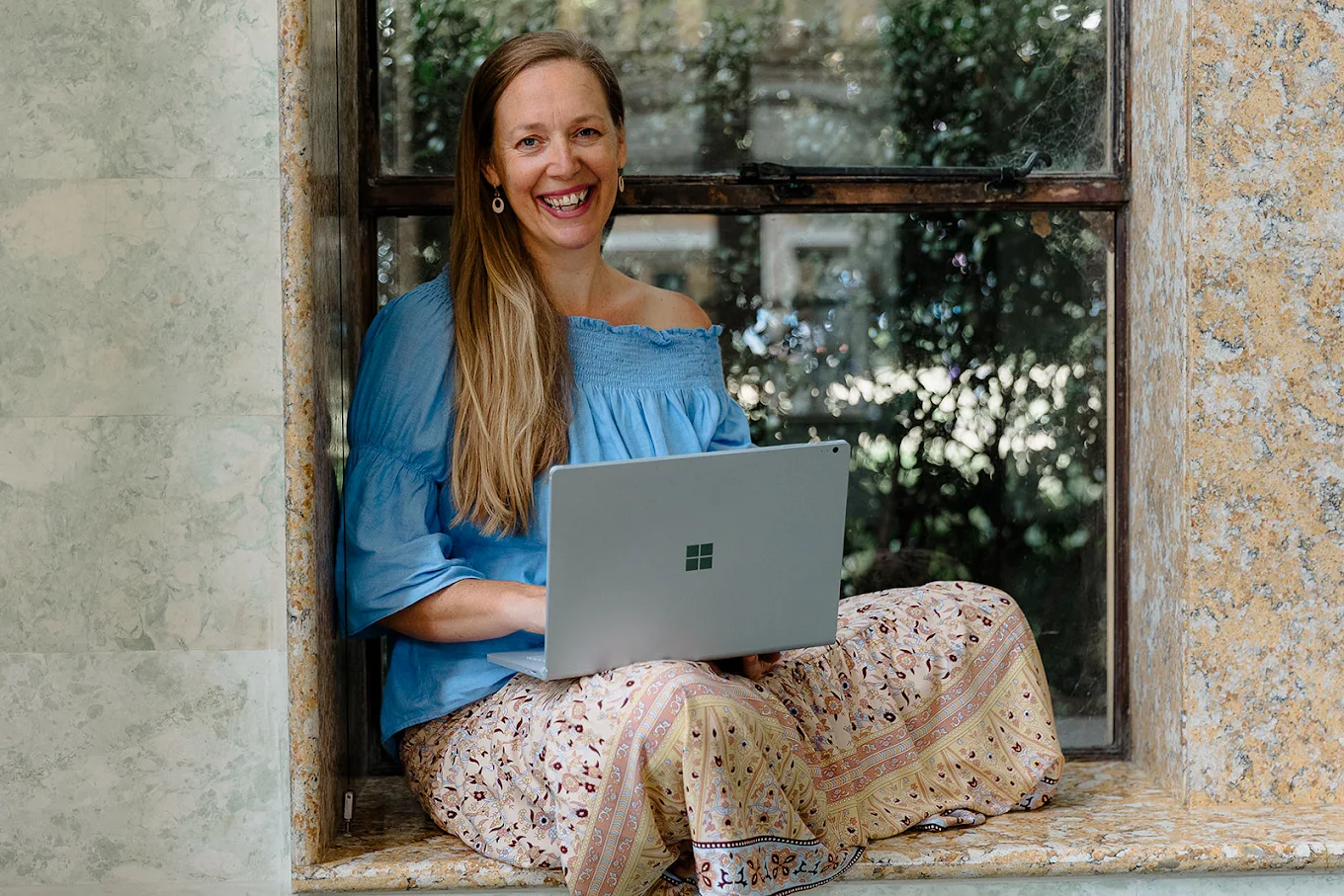 A woman sitting on a windowsill with a laptop, smiling at the camera.