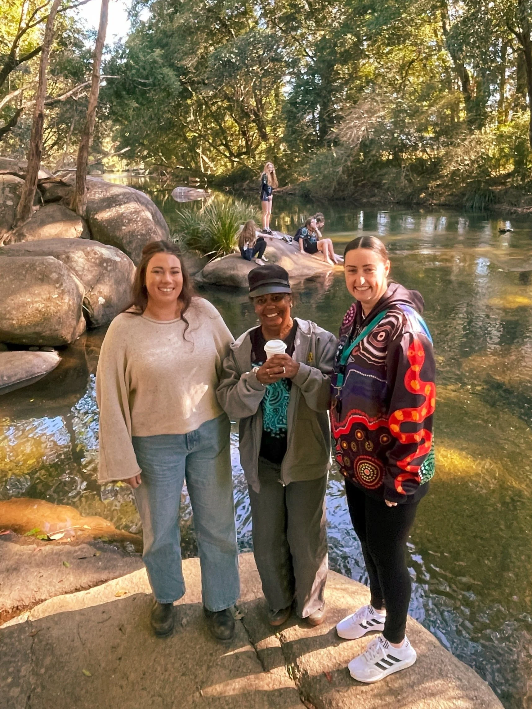 Three women standing on a rock by a river with trees in the background. Two women are smiling at the camera, one holds a coffee cup, and others are sitting or standing on rocks near the water.