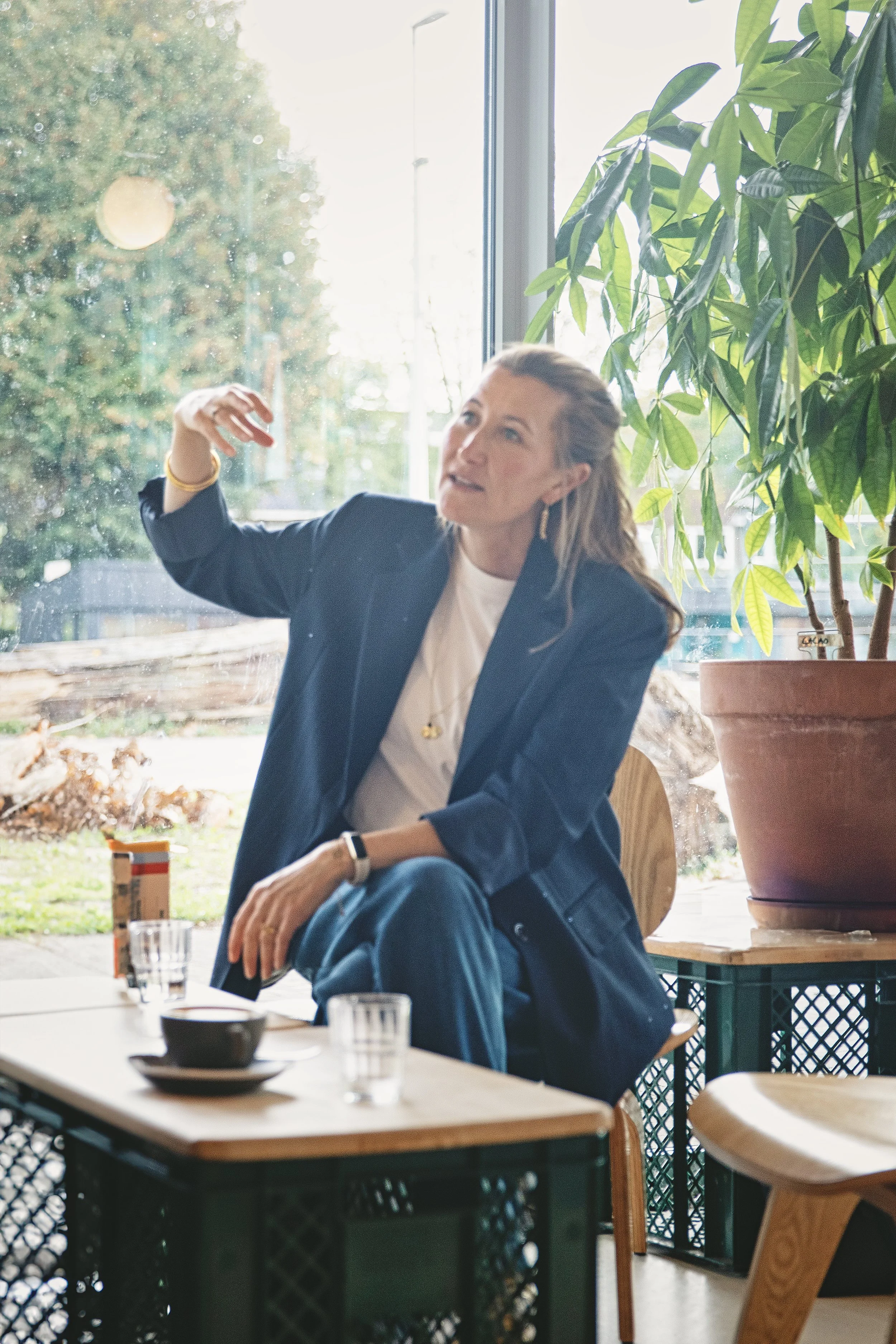 Vrouw in een kantoor of café, zittend op een houten stoel naast een tafel, die uitlegt of presenteert met handgebaren, grote planten en glasramen op de achtergrond.