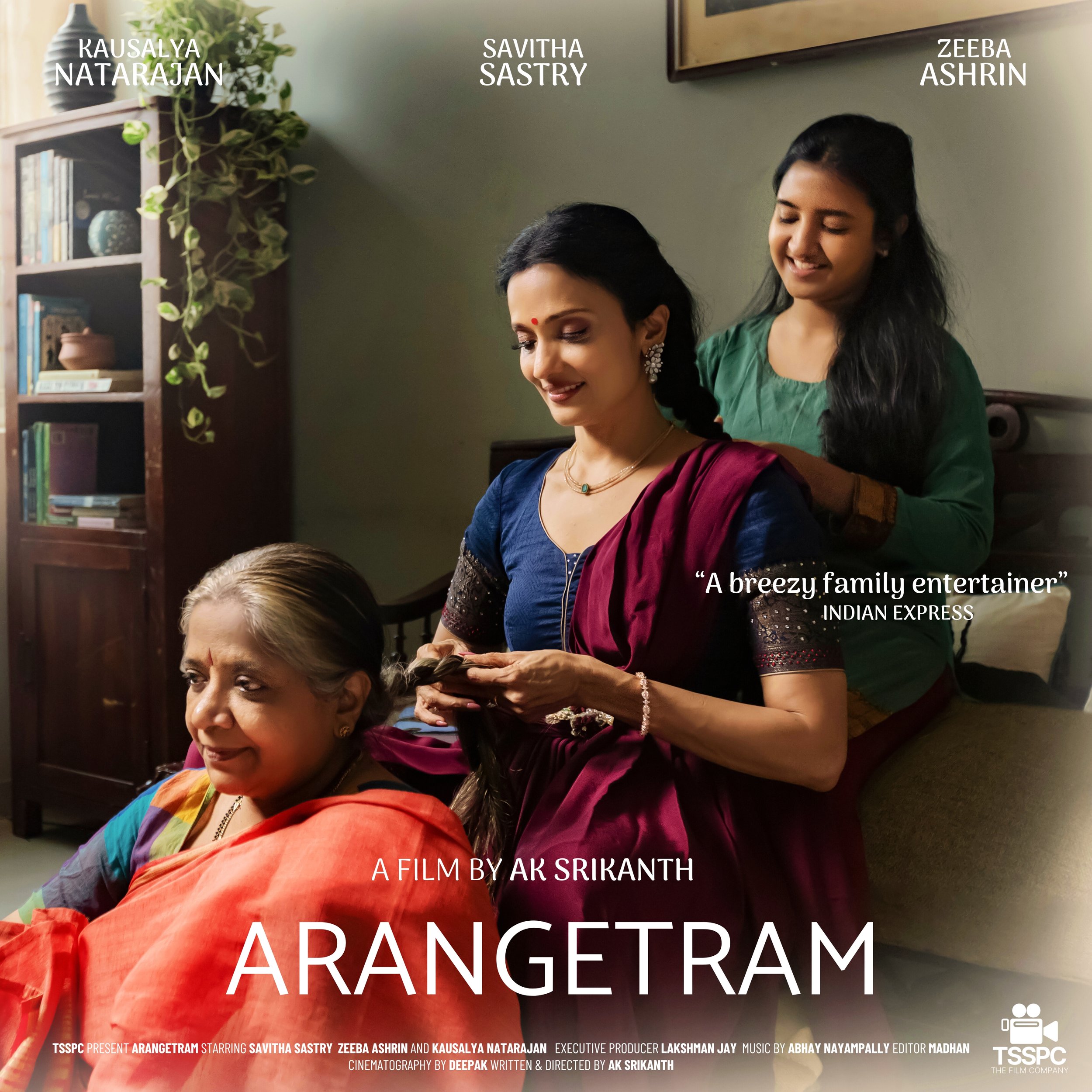 Three women, an elderly woman sitting on the floor and two younger women standing behind her, with one of the younger women combing the elderly woman's hair in a well-lit room with a bookshelf and window in the background. Savitha Sastry in Arangetra