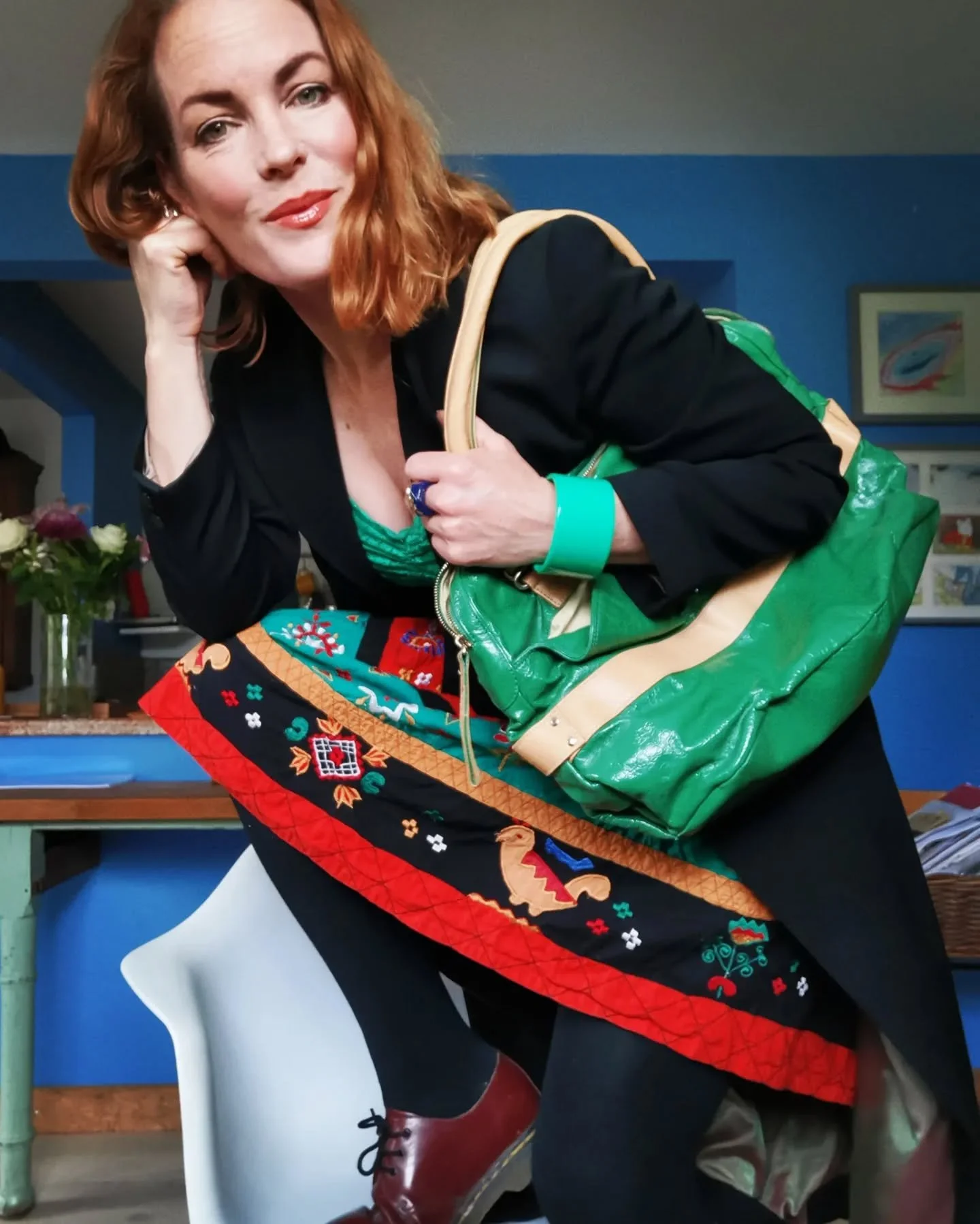 A woman with red hair and fair skin leaning forward, holding a green bag with yellow straps, wearing a black blazer, a colorful skirt, black tights, and red shoes, inside a room with blue walls and various decorations.