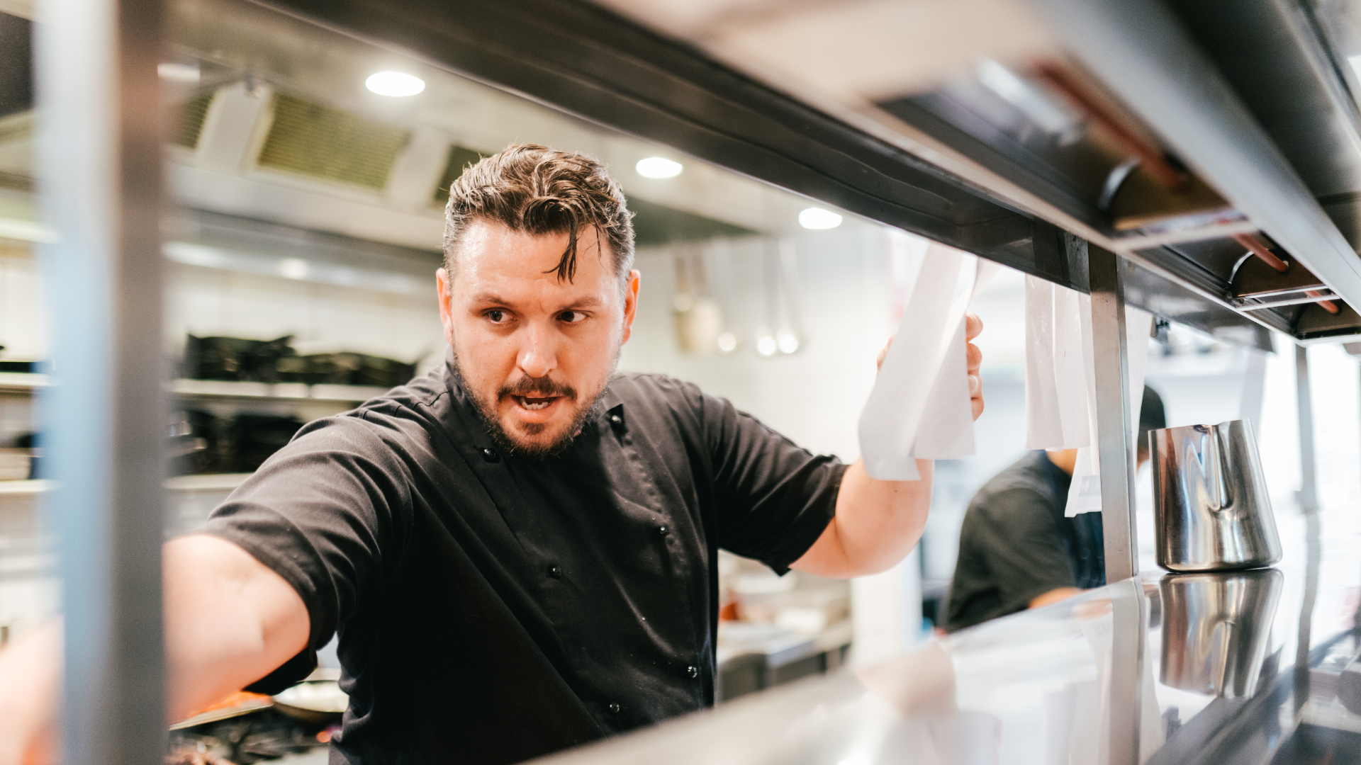A man with a beard and dark hair working in a restaurant kitchen, possibly a chef, leaning over a counter while looking intently at something outside the frame.