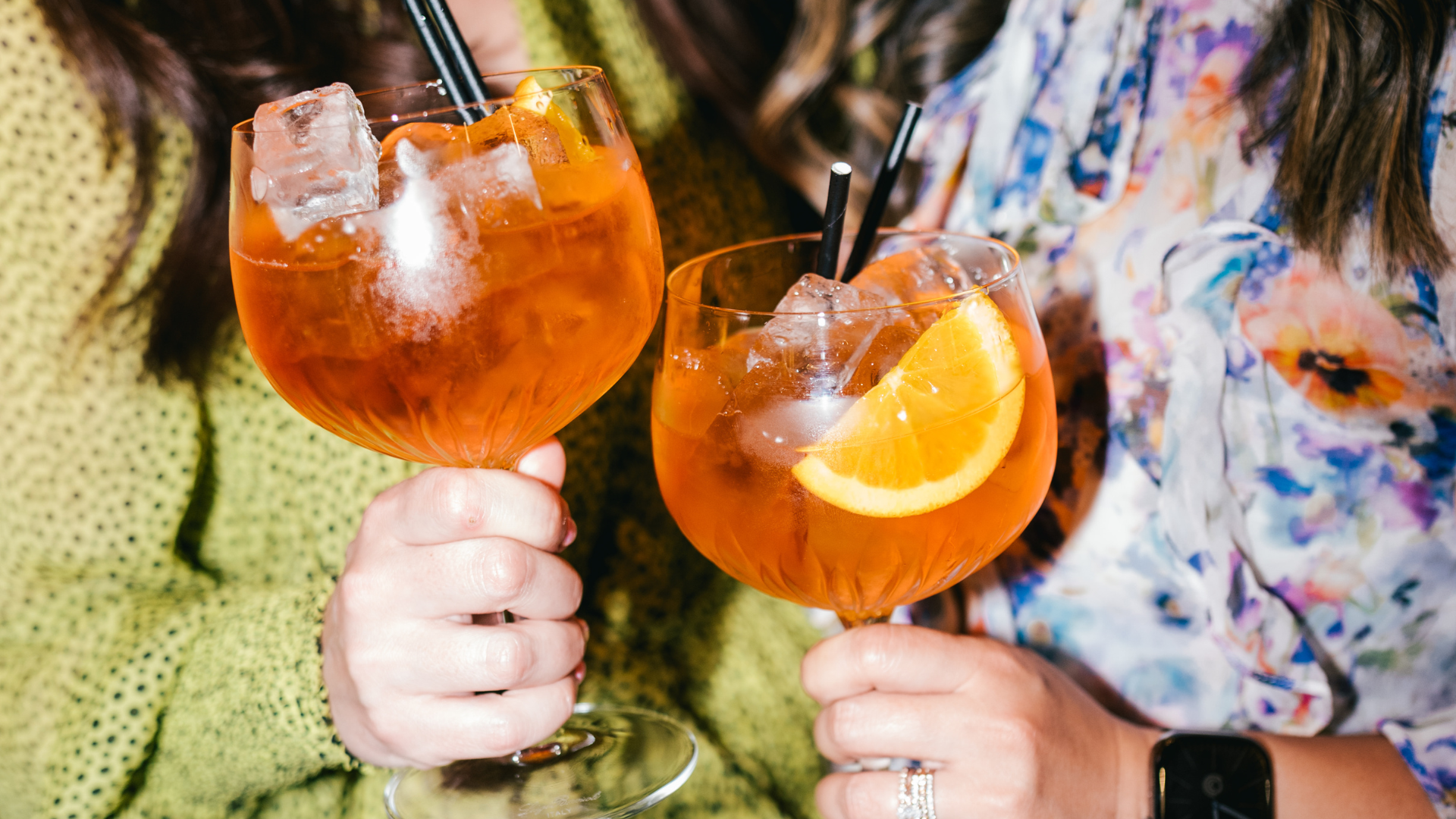 Two women holding large glasses of orange-colored cocktail garnished with orange slices, ice cubes, and black straws.