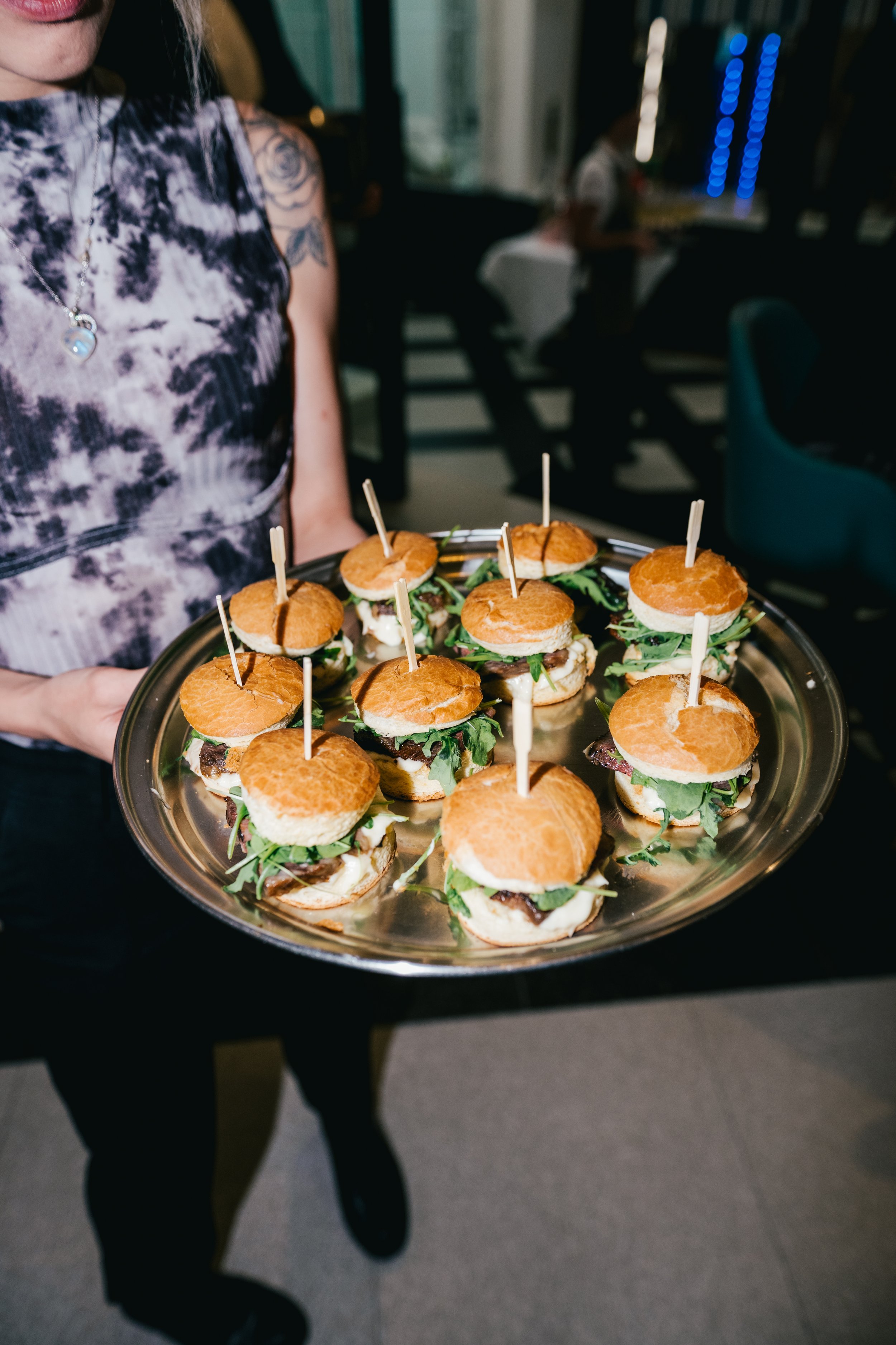 A person dressed in black holding a large tray of mini sandwiches with bun tops secured by skewers, in a dimly lit indoor setting, with a woman in a tie-dye dress and silver necklace visible in the background.