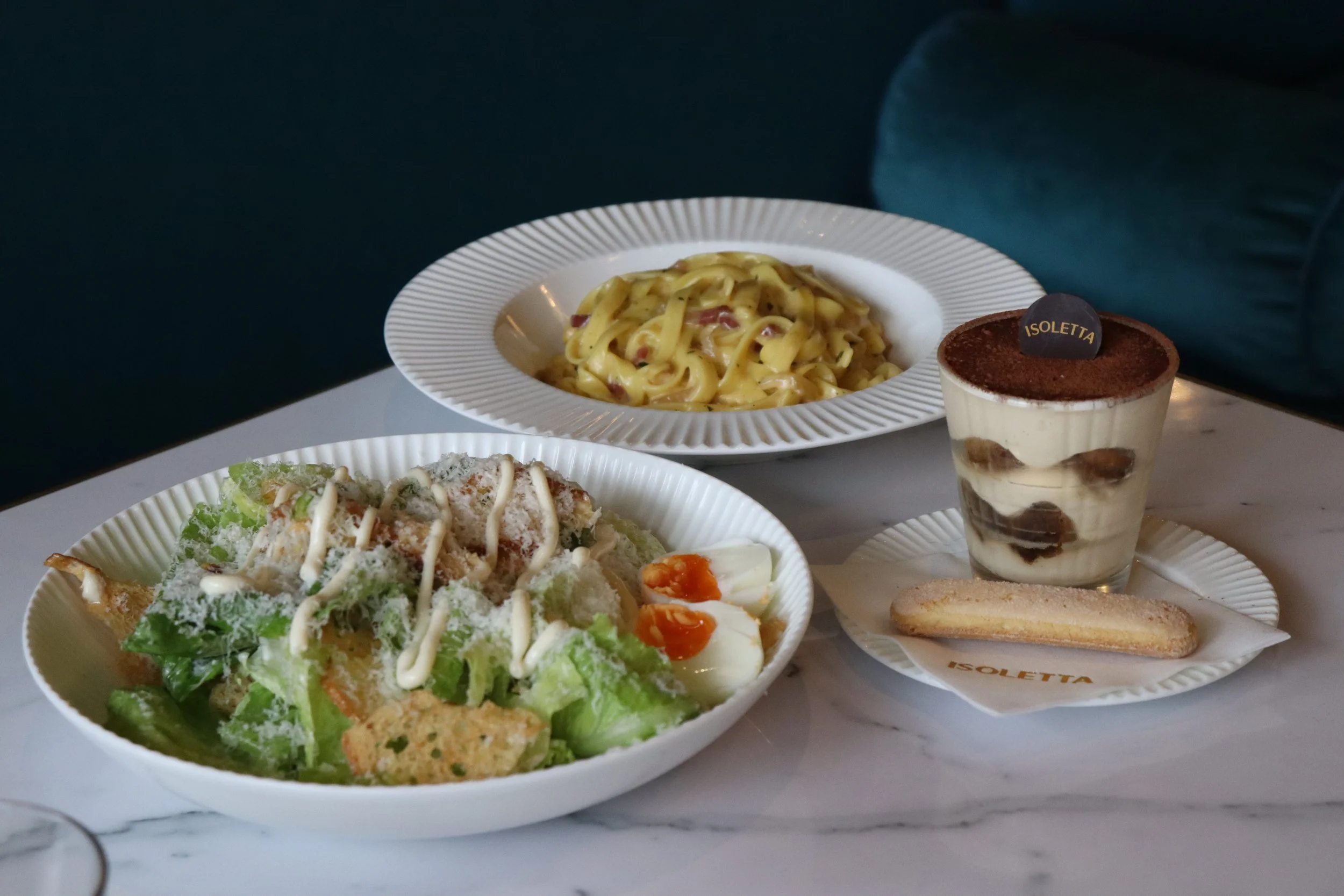 A plate of linguine pasta with creamy sauce and pieces of meat, and a salad with lettuce, grated cheese, and sliced boiled eggs, along with a dessert of tiramisu and a ladyfinger cookie on a white marble table.