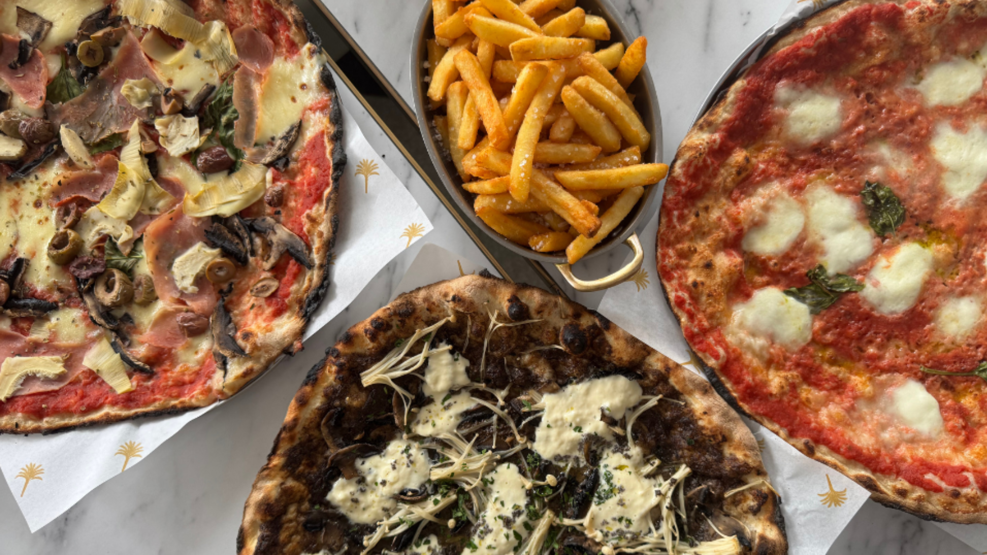 Three pizzas topped with various ingredients, a side of French fries, all on white paper with a palm tree pattern, set on a marble surface.