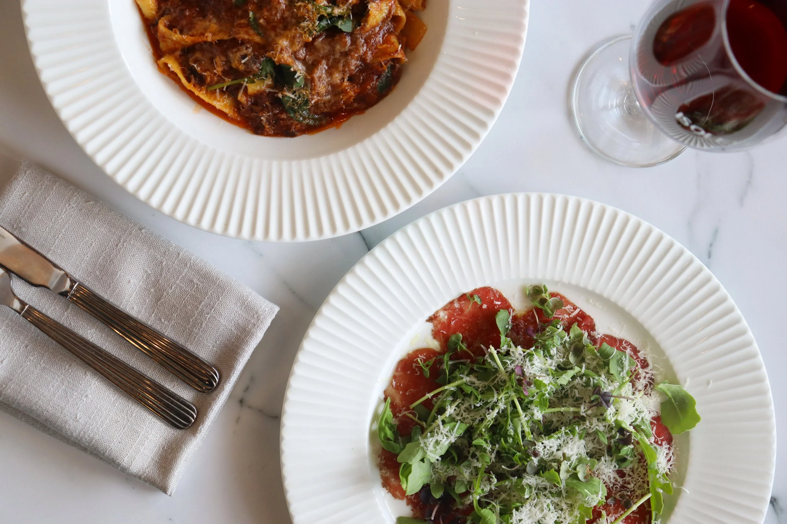 A close-up of two white, ridged, round plates with different dishes on a white marble table. One dish appears to be lasagna with sauce and herbs, and the other is a salad with greens, grated cheese, and tomato slices. There is a folded beige napkin w