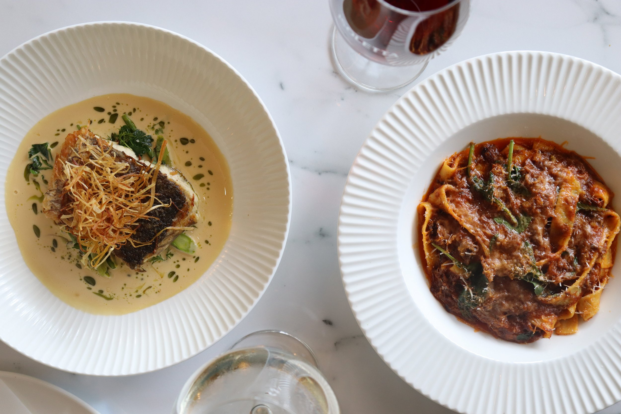 Two white bowls of pasta sit on a white marble table, with a glass of red wine and a partially visible glass of white wine nearby.