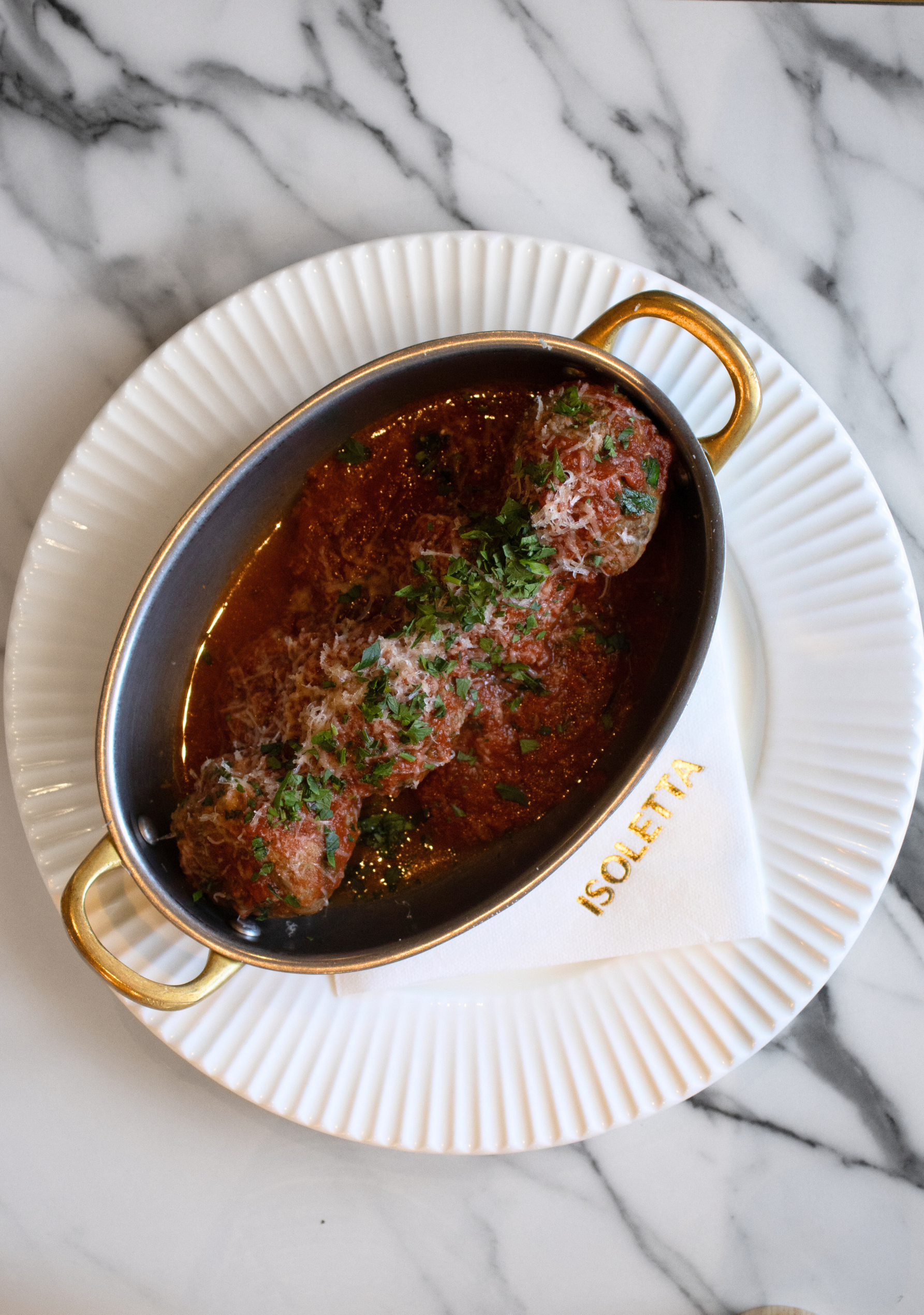 Two meatballs served in a small metal dish with tomato sauce, garnished with chopped parsley and grated Parmesan cheese, placed on a white plate with a gold-rimmed napkin, on a marble surface.