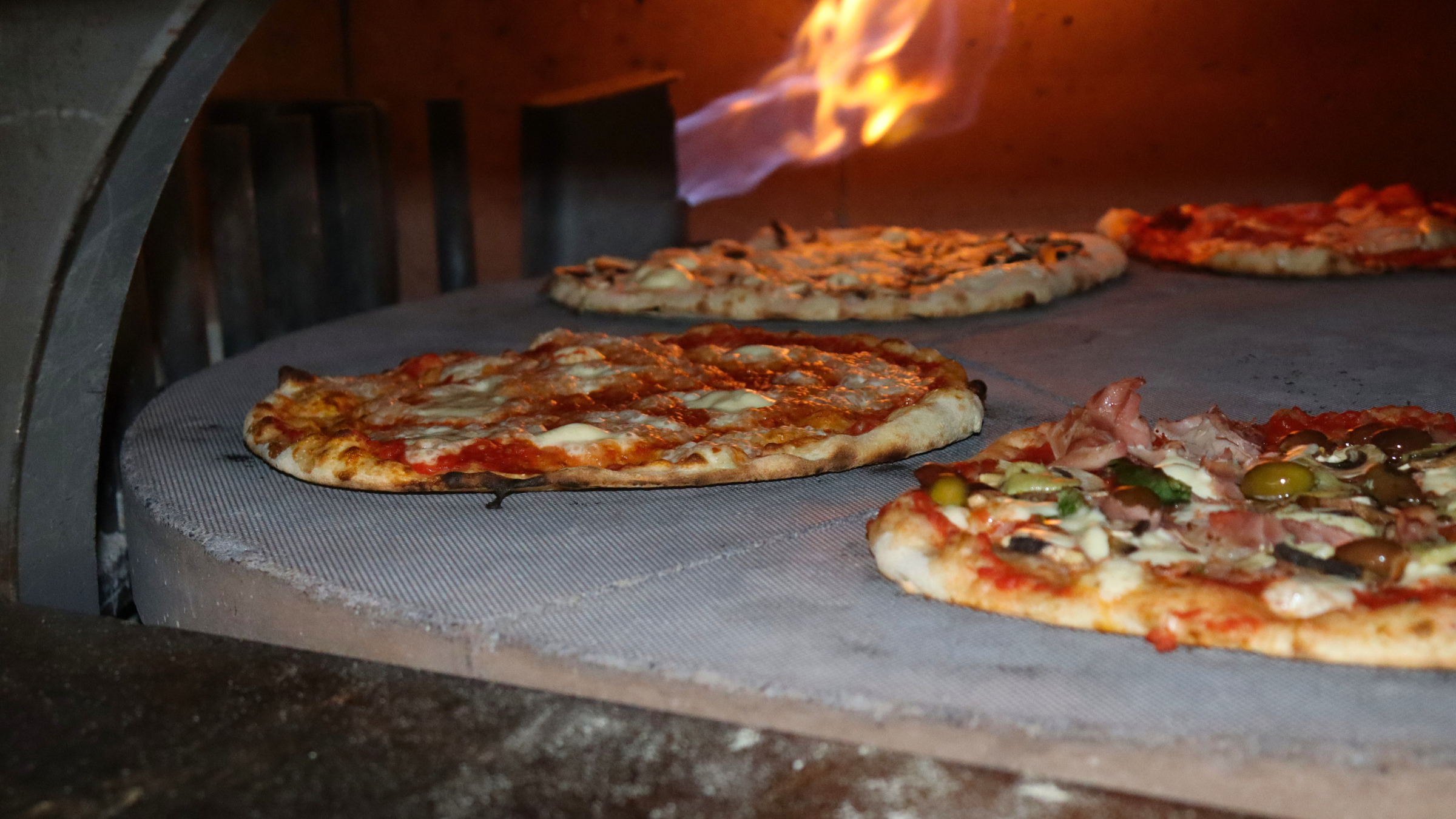 Four pizzas baking inside a wood-fired oven, with flames visible in the background.