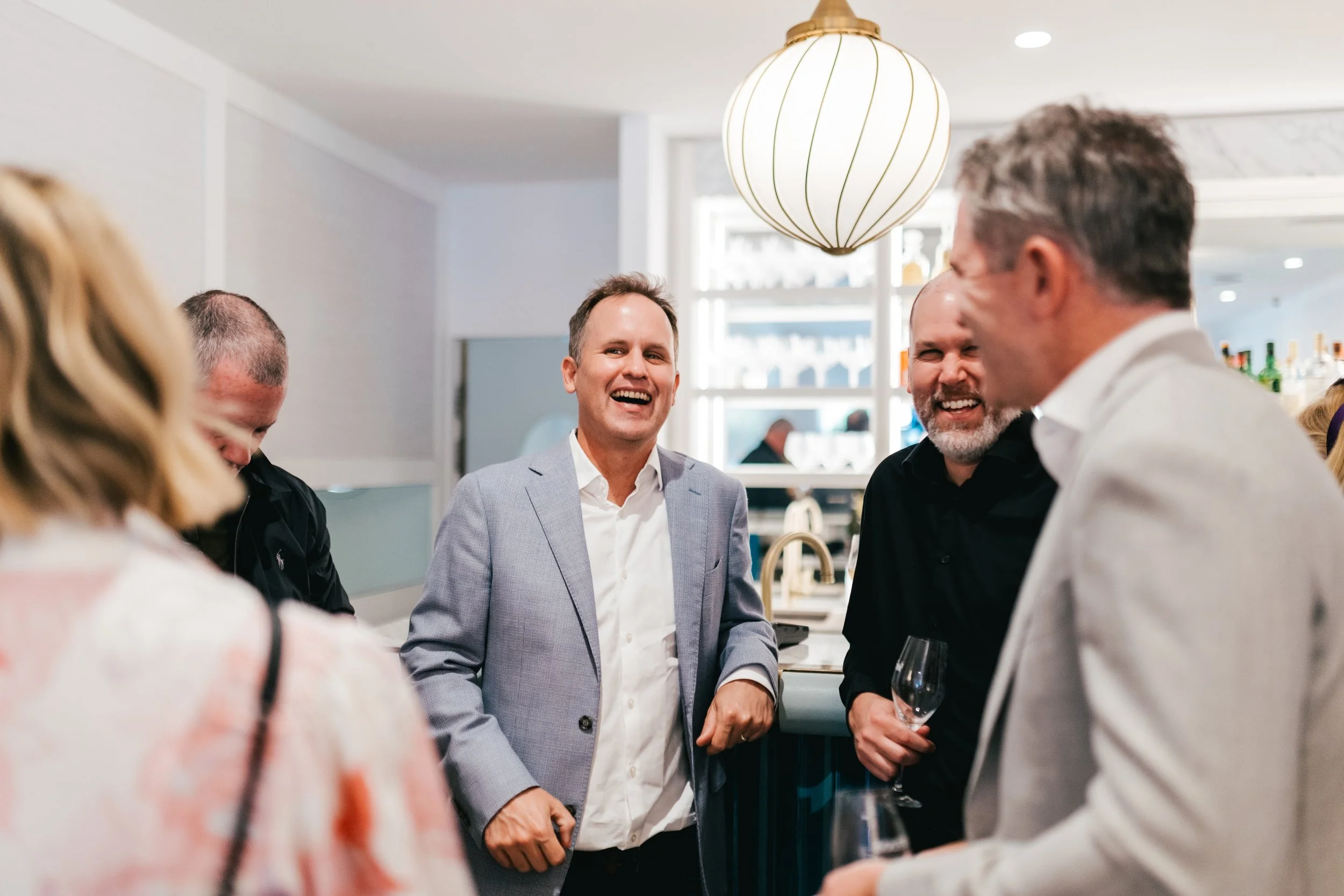 Group of people socializing inside a modern room, with one man in a light gray suit smiling and others holding wine glasses.