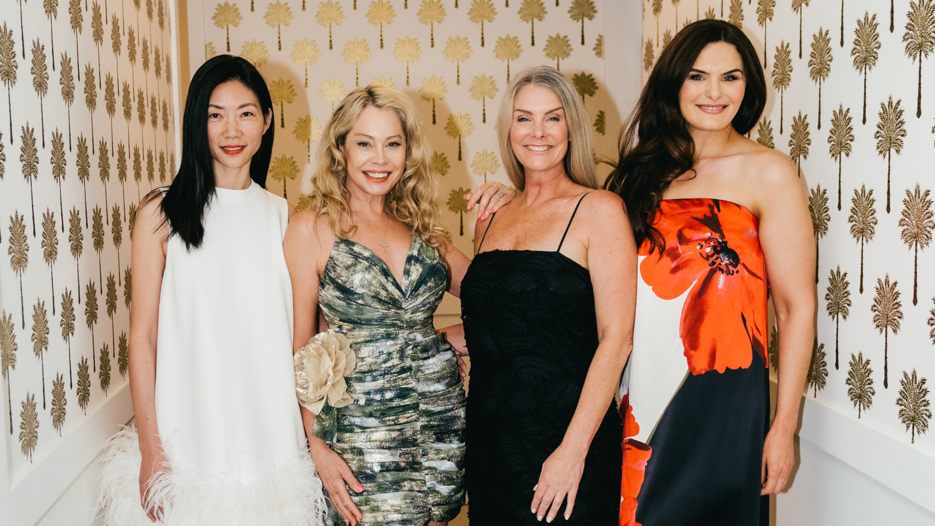 Four women pose together indoors, smiling, with patterned wallpaper featuring palm trees in the background. They are dressed in formal wear and standing close.