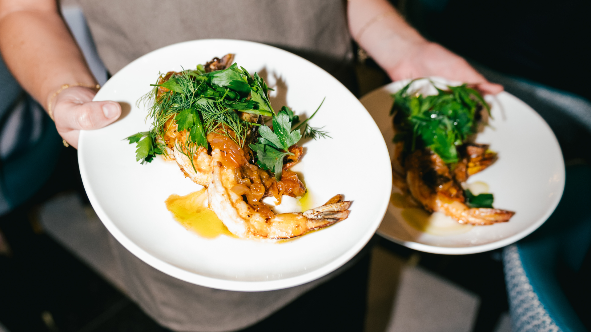 A person holding two plates with grilled shrimp topped with herbs and sauce, garnished with fresh parsley and dill.