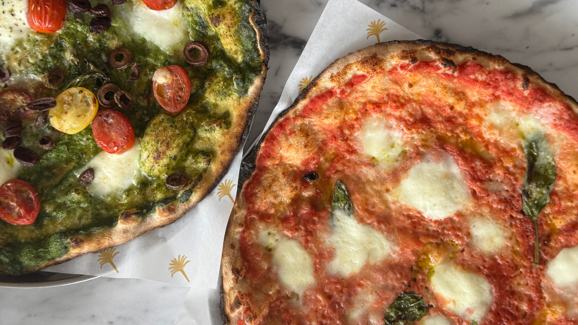 Two pizzas on a marble surface, one with cherry tomatoes, olives, and green sauce, and the other with tomato sauce, mozzarella, and basil.